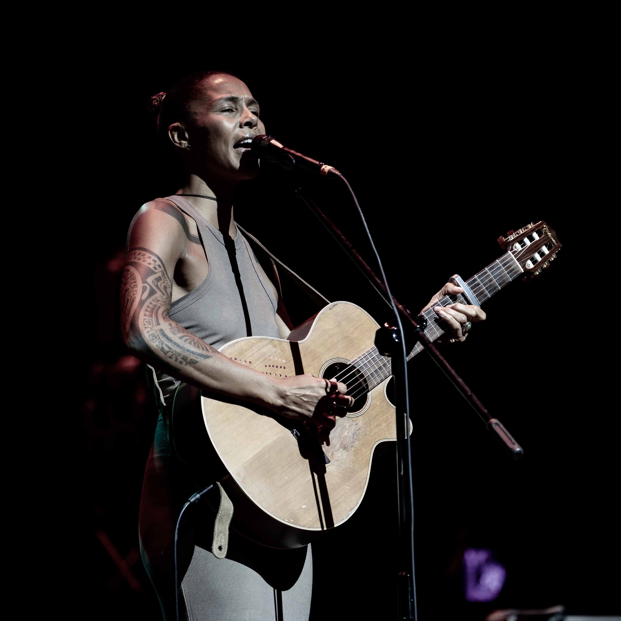 Koncertowe ujęcie: wokalistka z tatuażem na ramieniu śpiewa, grając na gitarze akustycznej przy mikrofonie na tle ciemnej sceny, oświetlona punktowym światłem.
