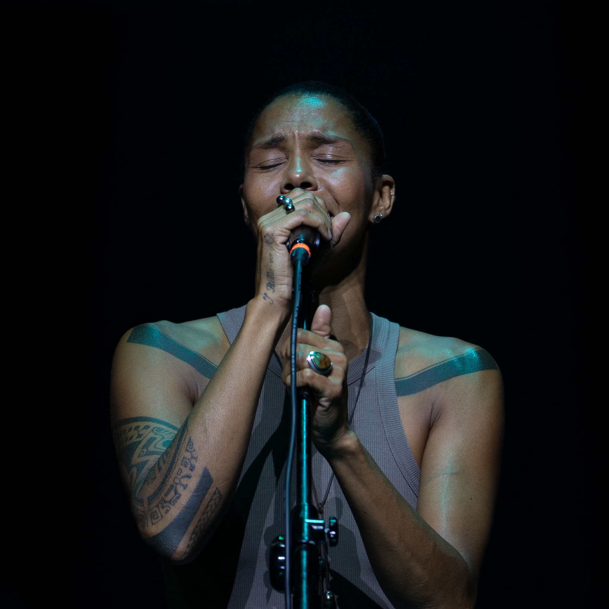 Artystka z tatuażami na ramionach, śpiewa z zamkniętymi oczami do mikrofonu na czarnym tle, oświetlona punktowym światłem. Widoczne pierścionki na dłoniach.