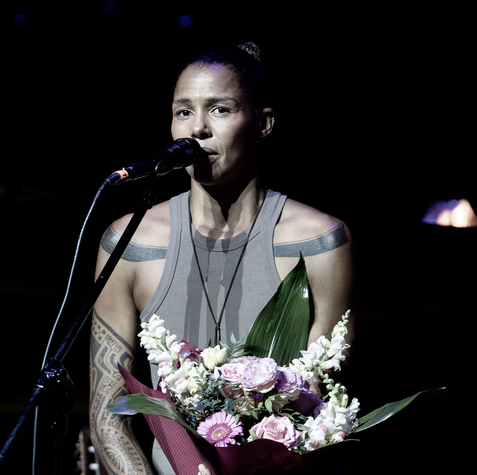 Piosenkarka z tatuażami na ramionach, śpiewa do mikrofonu, trzymając bukiet kwiatów na scenie w ciemnym oświetleniu. Koncertowy kadr.