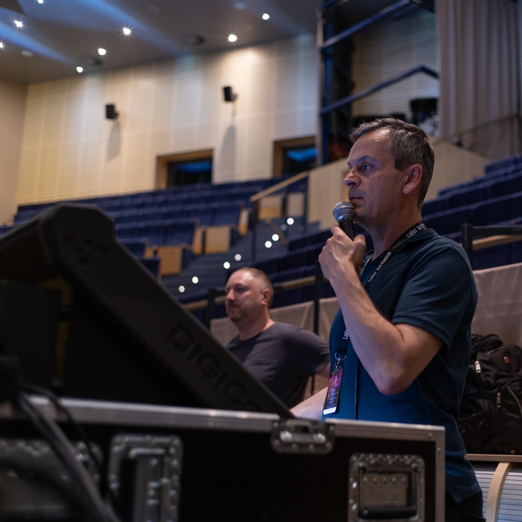 Mężczyzna z mikrofonem na tle widowni i sprzętu nagłośnieniowego. Ujęcie z dołu, widoczny fragment sceny i oświetlenie. Konferencja lub koncert.