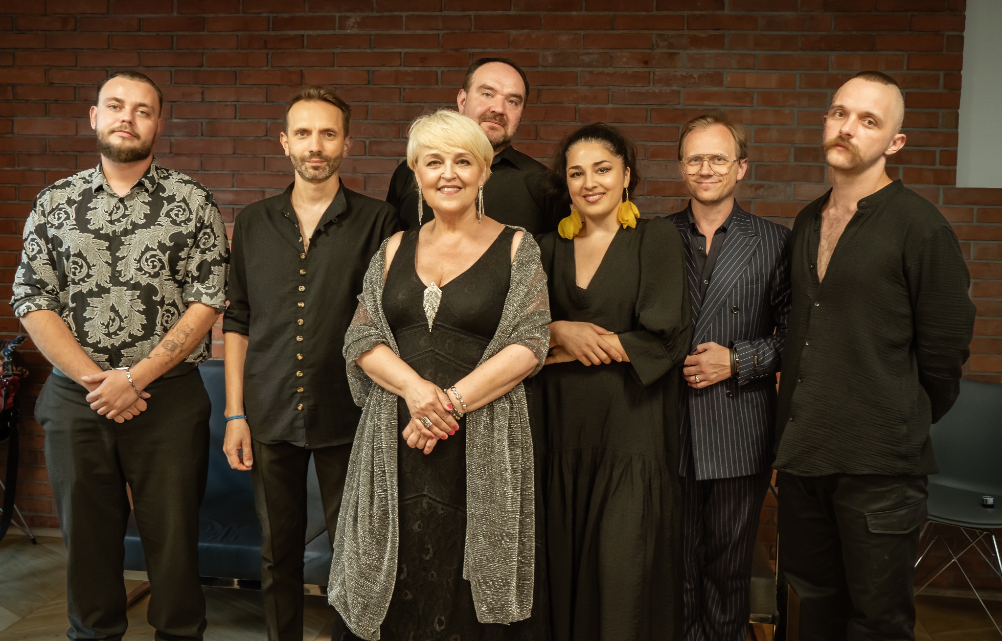 Grupa siedmiu osób w ciemnych strojach pozuje na tle ceglanej ściany. Kobieta w sukni wieczorowej na pierwszym planie. Ujęcie studyjne, oświetlenie równomierne.