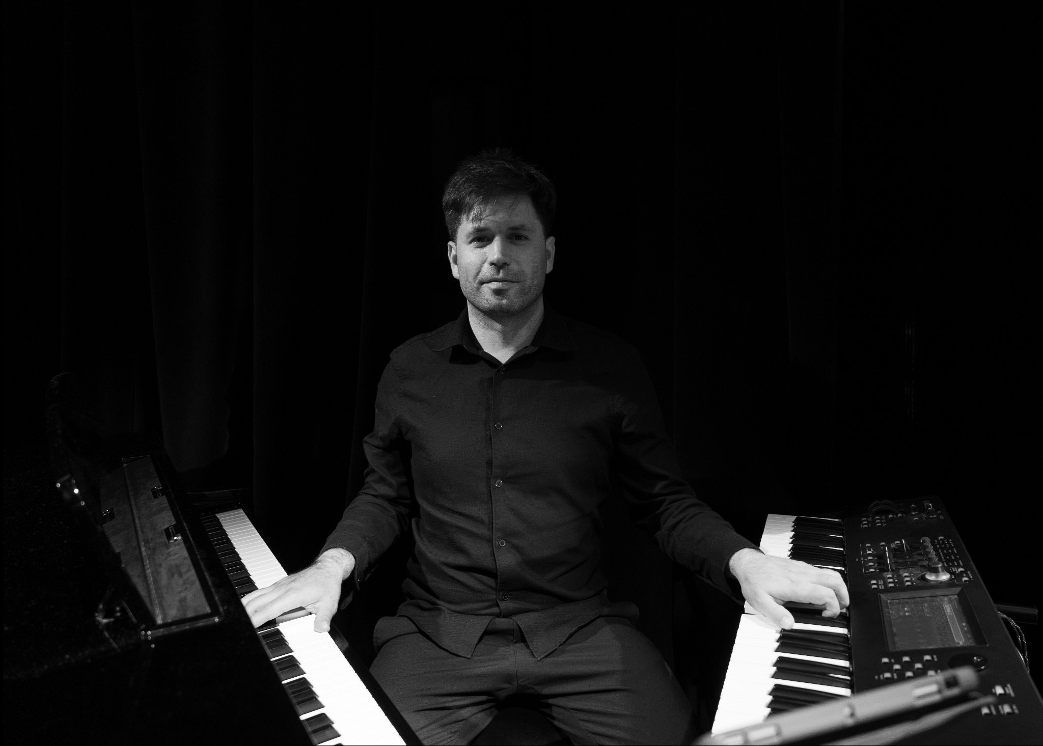 Czarno-białe zdjęcie muzyka siedzącego za dwoma keyboardami, z rękami na klawiszach, na ciemnym tle. Ujęcie centralne, skupione na twarzy i instrumentach.