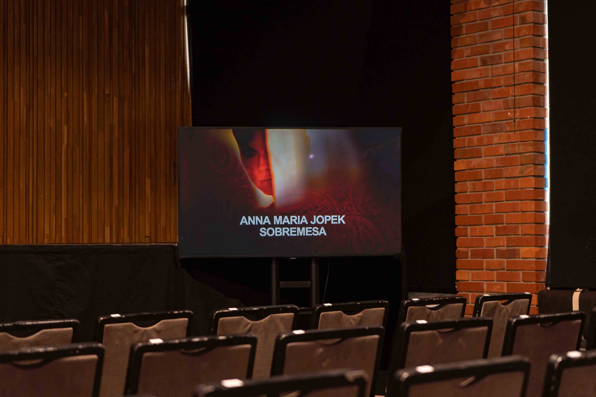 Sala koncertowa z rzędami krzeseł i ekranem wyświetlającym Annę Marię Jopek. Ceglana kolumna i drewniana ściana dodają charakteru wnętrzu. Scena gotowa na występ.