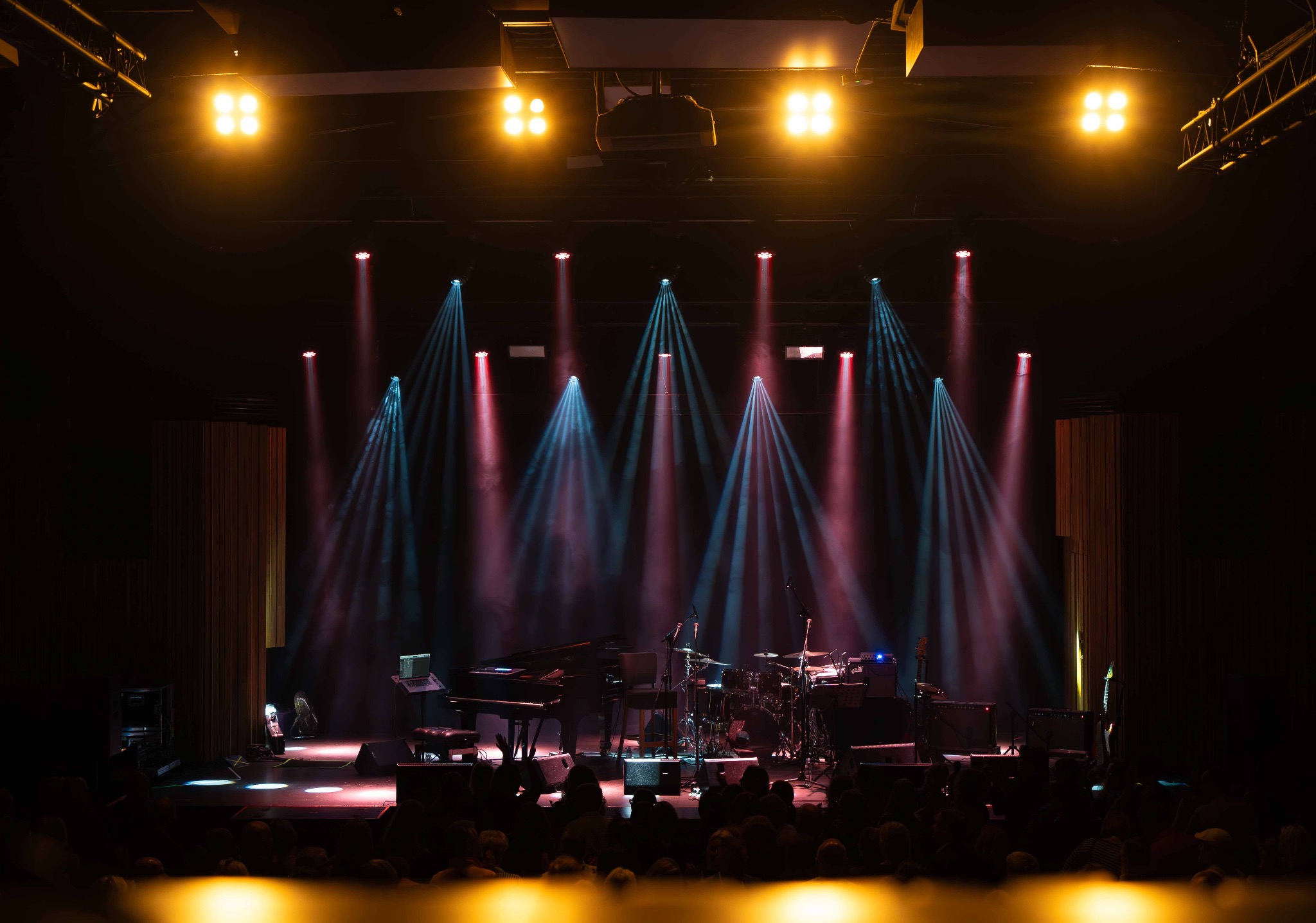 Scena koncertowa z instrumentami (pianino, perkusja) oświetlona reflektorami w kolorach żółtym, czerwonym i niebieskim. Widoczna publiczność w tle.
