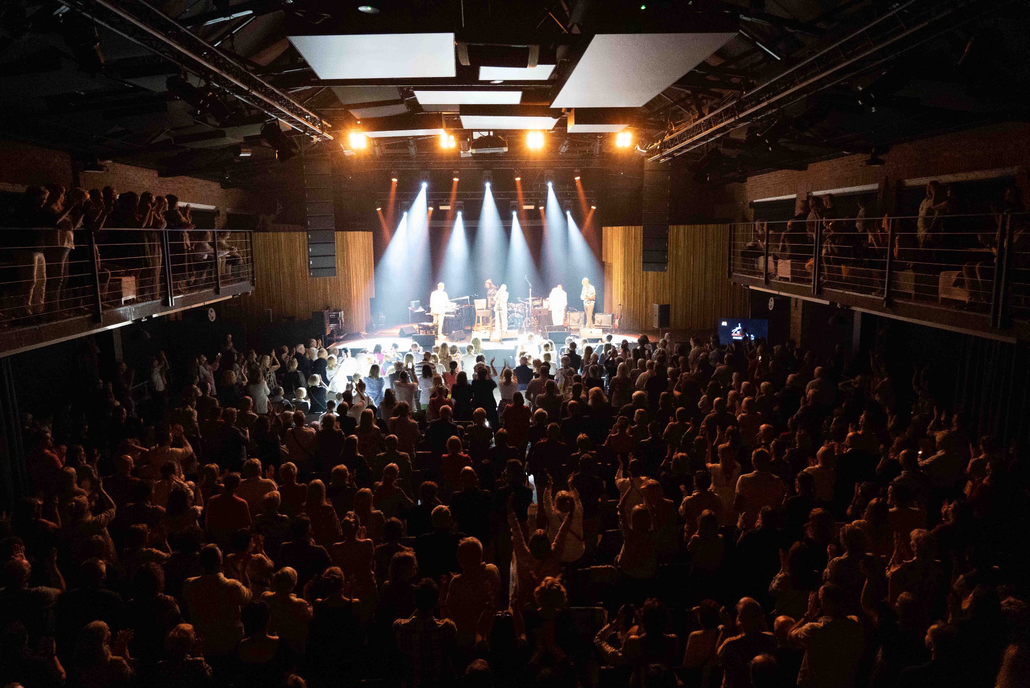 Koncert w Gdańsku: zespół na scenie w blasku reflektorów, tłum publiczności w ciemności. Ujęcie z góry, widok na całą salę koncertową.