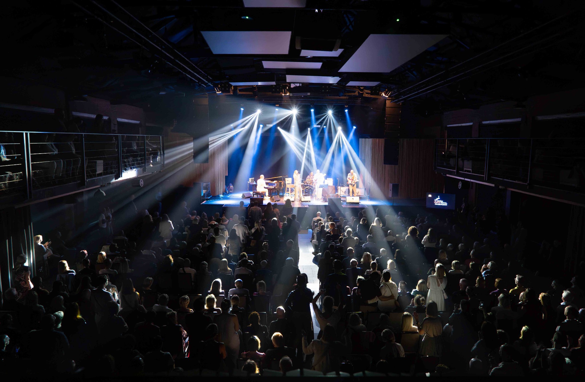Koncert z widokiem na scenę i publiczność w ciemnym pomieszczeniu, oświetloną reflektorami. Zespół muzyczny na scenie. Widok z góry na widownię.