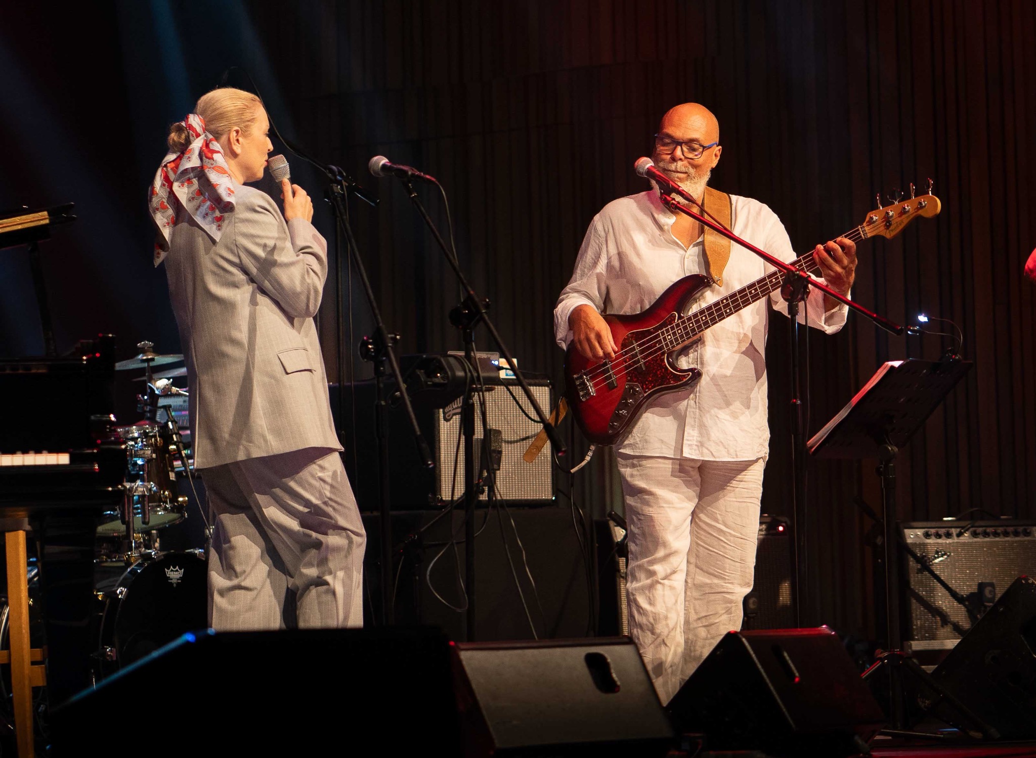 Duet muzyczny na scenie: kobieta w szarym garniturze śpiewa do mikrofonu, obok mężczyzna gra na basie w białym stroju. W tle instrumenty i oświetlenie sceniczne.