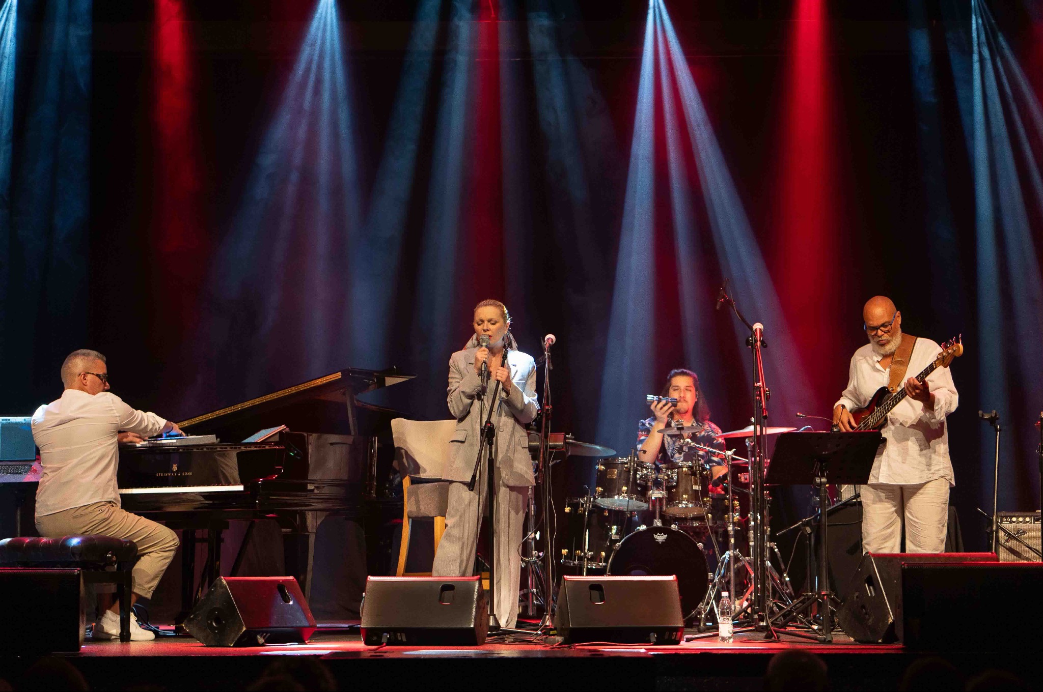 Zespół muzyczny na scenie podczas koncertu: wokalistka w szarym garniturze, pianista, perkusista i basista. Czerwono-niebieskie światła sceniczne w tle.