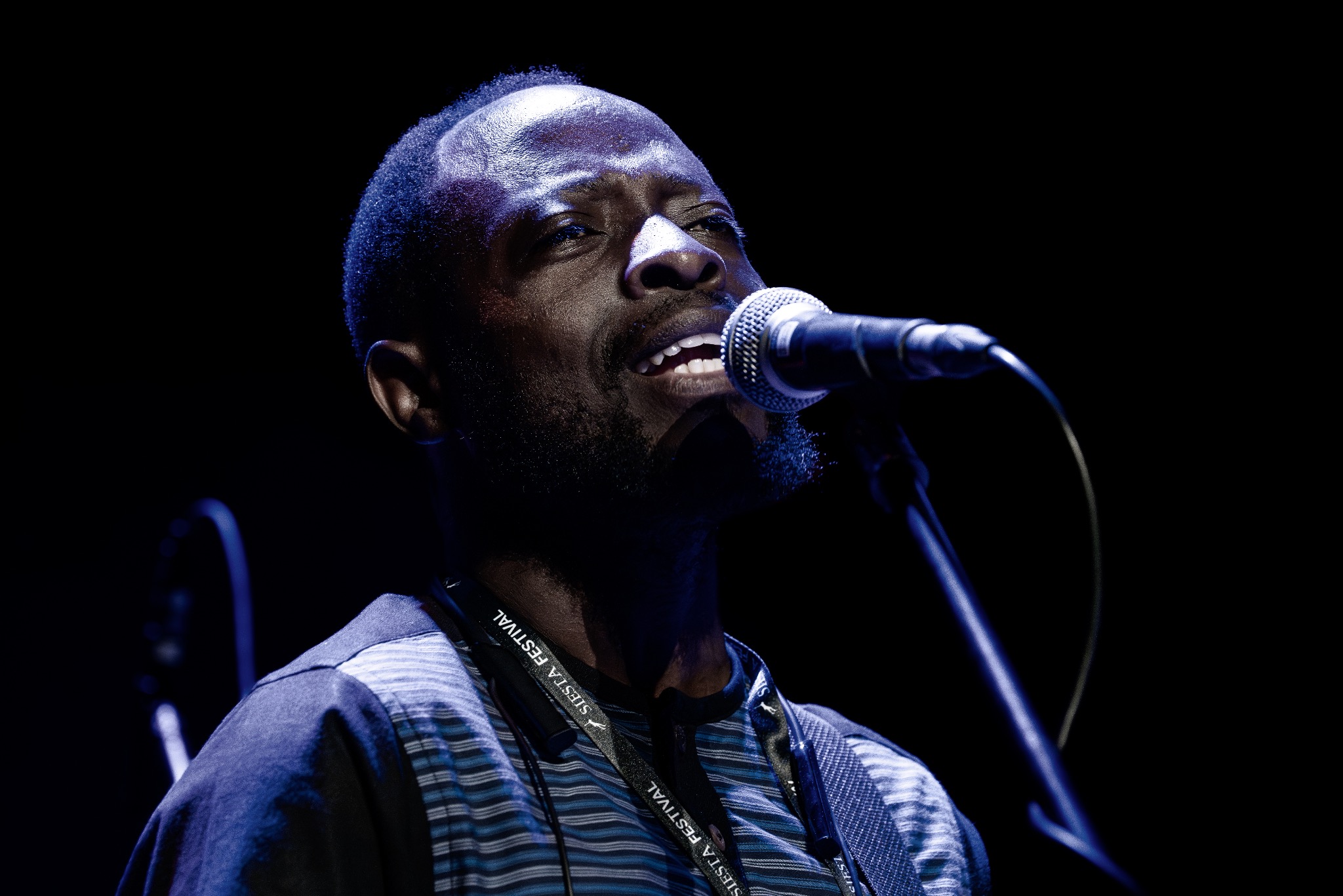 Portret wokalisty z mikrofonem na scenie, oświetlonego niebieskim światłem. Widoczny fragment koszuli w paski i smycz z napisem 'Siesta Festival'.