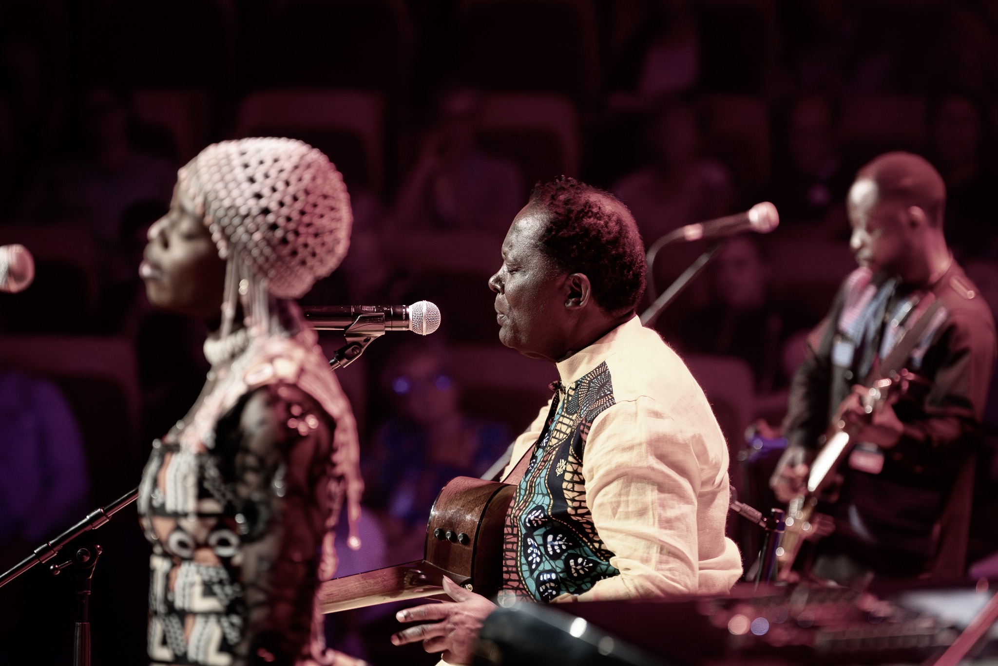 Koncertowy kadr: Muzyk gra na gitarze, obok wokalistka w etnicznym stroju, w tle gitarzysta. Scena oświetlona w odcieniach czerwieni i fioletu, widoczna publiczność.