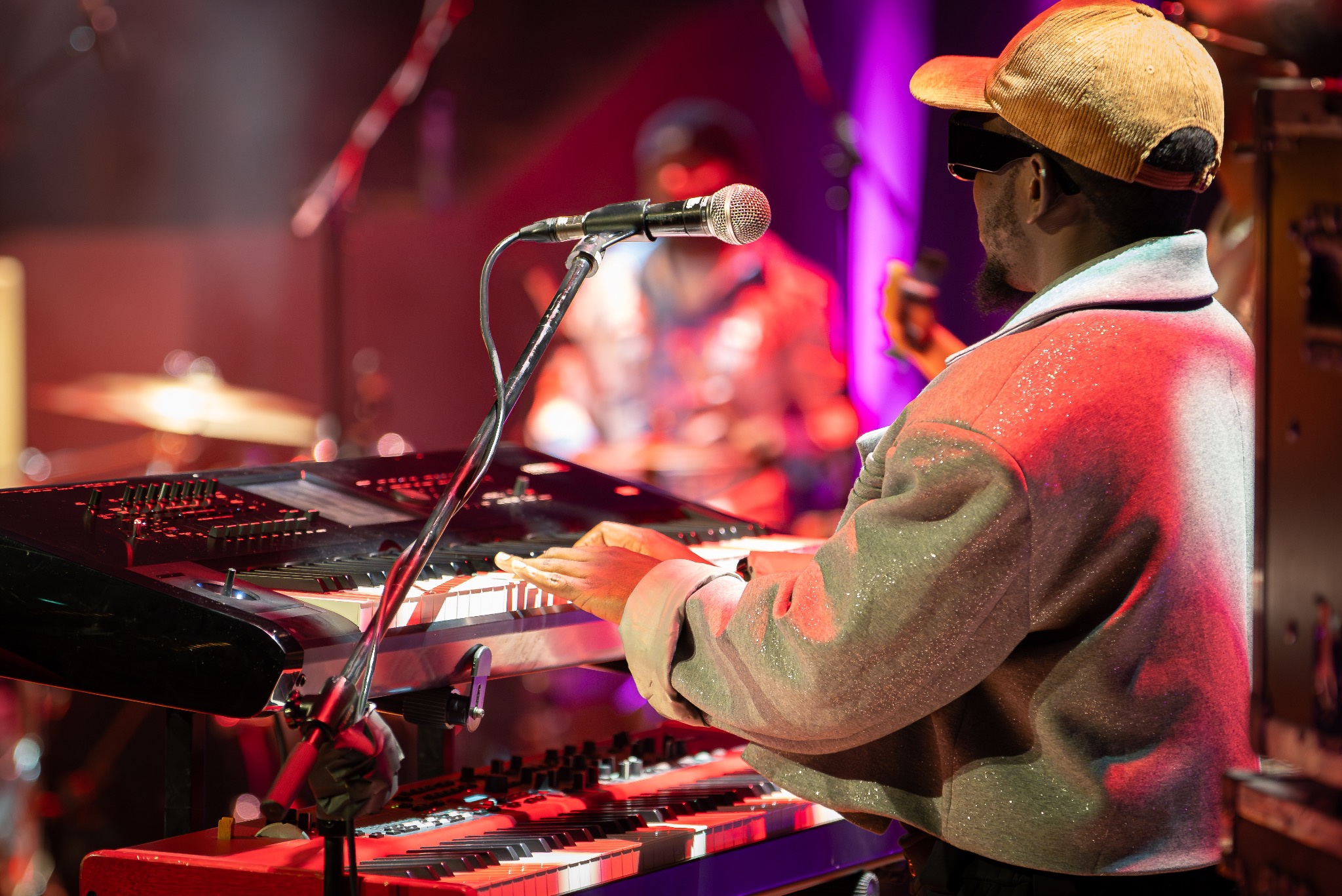 Artysta w błyszczącej marynarce gra na keyboardzie podczas koncertu, mikrofon na statywie, perkusja w tle, oświetlenie sceniczne.