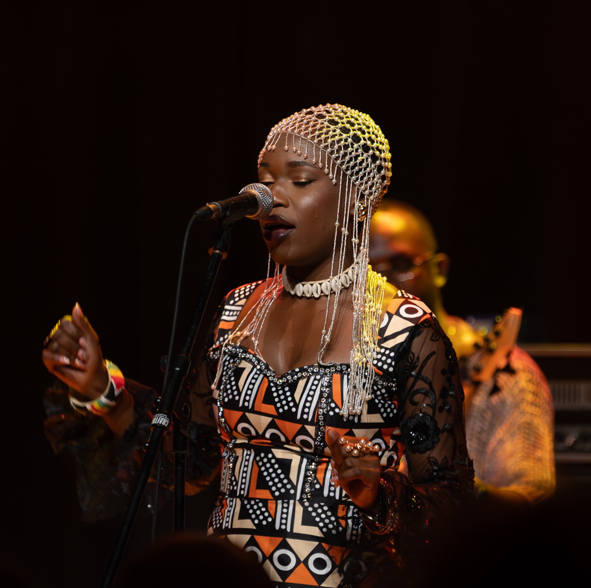 Piosenkarka w etnicznym stroju i nakryciu głowy śpiewa do mikrofonu na scenie podczas koncertu. W tle widoczny muzyk z instrumentem.