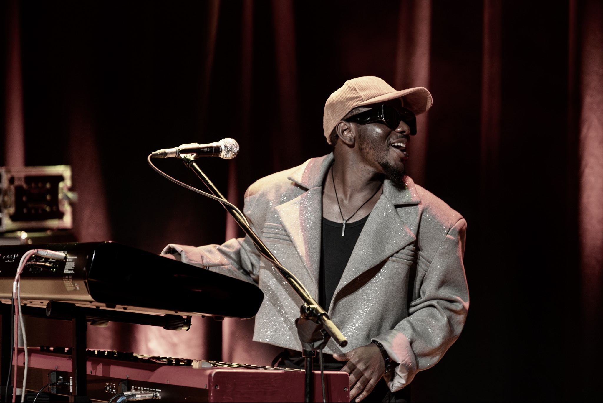 Uśmiechnięty muzyk w okularach i czapce gra na keyboardzie podczas koncertu. Widoczny mikrofon i oświetlenie sceniczne. Ciemne tło z czerwonymi akcentami.