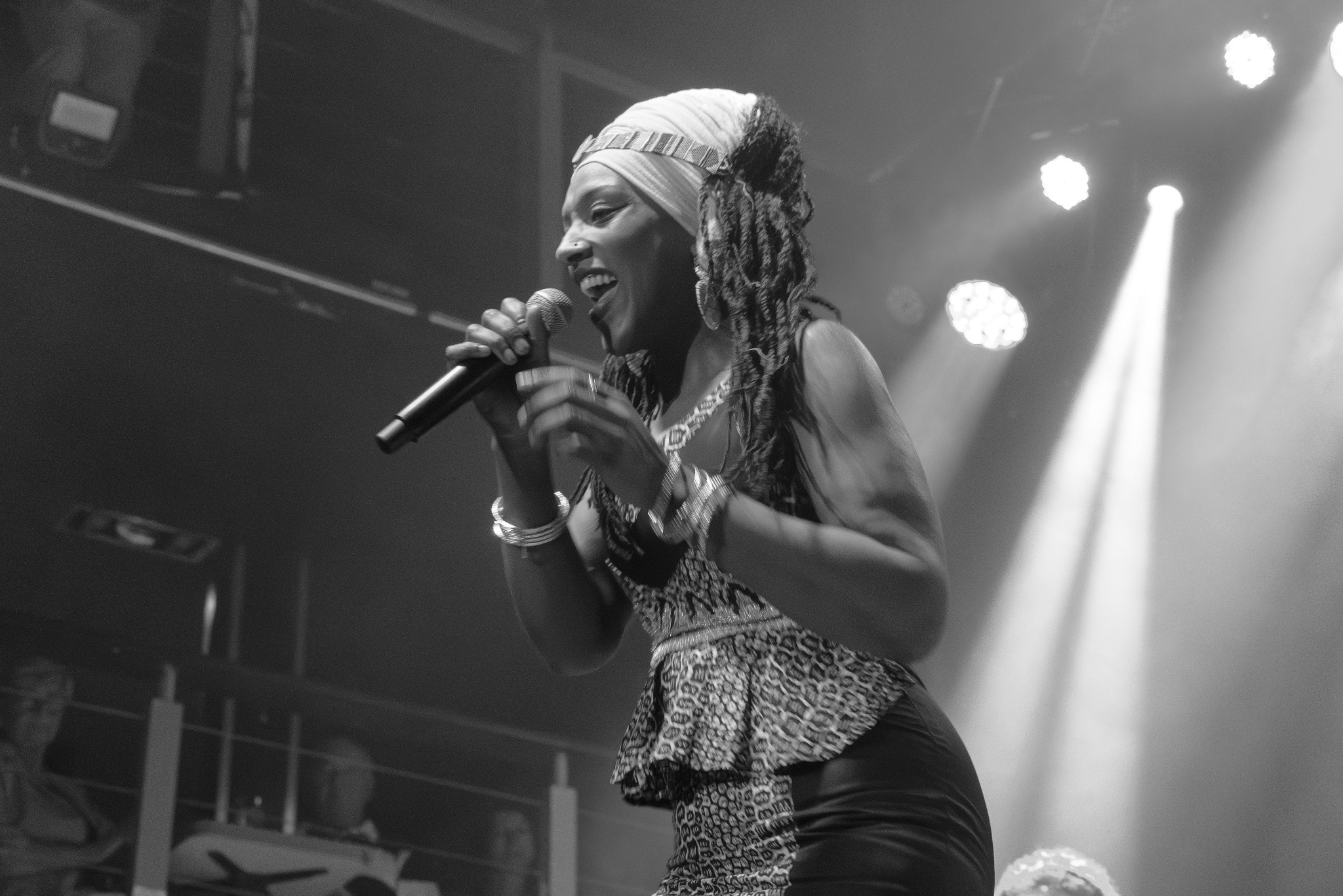 Czarno-białe zdjęcie piosenkarki z dredami, w bluzie w panterkę, śpiewającej do mikrofonu na scenie podczas koncertu, oświetlonej reflektorami.