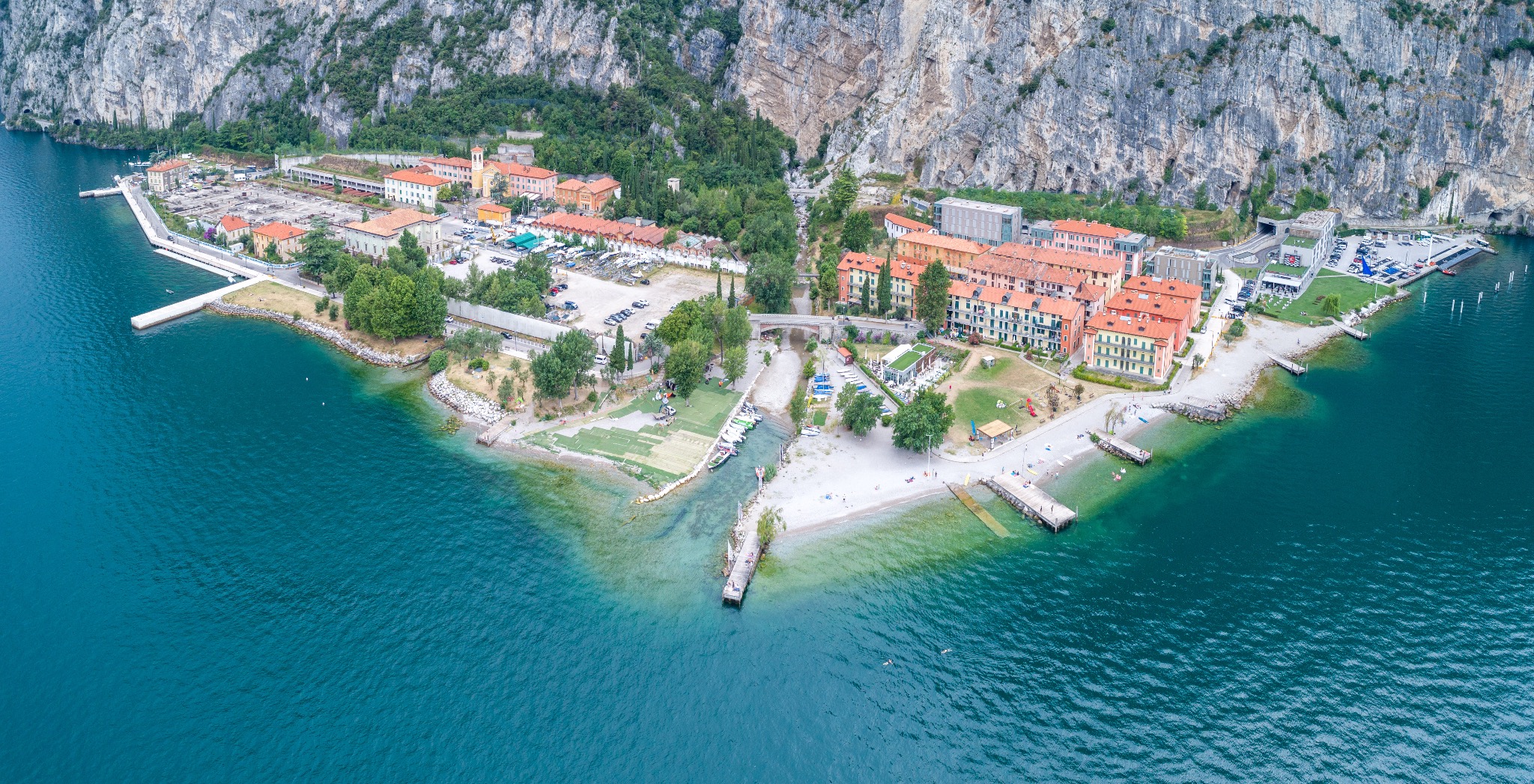 Panoramiczny widok z drona na miasteczko nad jeziorem Garda, otoczone górami. Widoczne budynki z czerwonymi dachami, plaża, przystań i turkusowa woda.