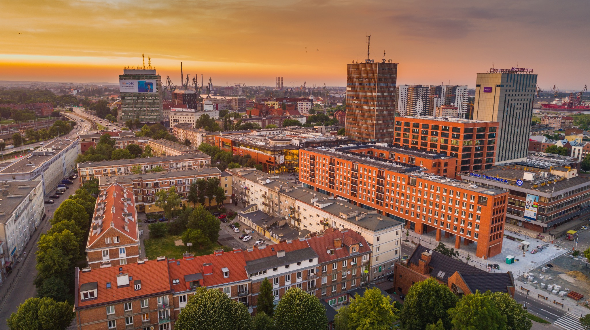 Panoramiczny widok z drona na Gdańsk o zachodzie słońca. Widoczne budynki mieszkalne, biurowce, dźwigi portowe i pomarańczowe niebo.