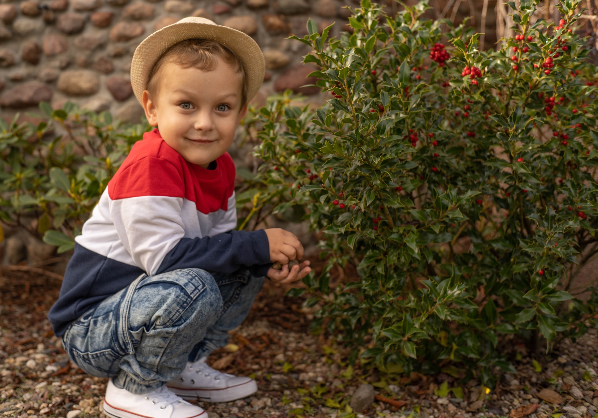 Uśmiechnięty chłopiec w kapeluszu kuca obok krzewu z czerwonymi owocami. Dziecko ma na sobie bluzę w kolorach czerwieni, bieli i granatu oraz dżinsy.