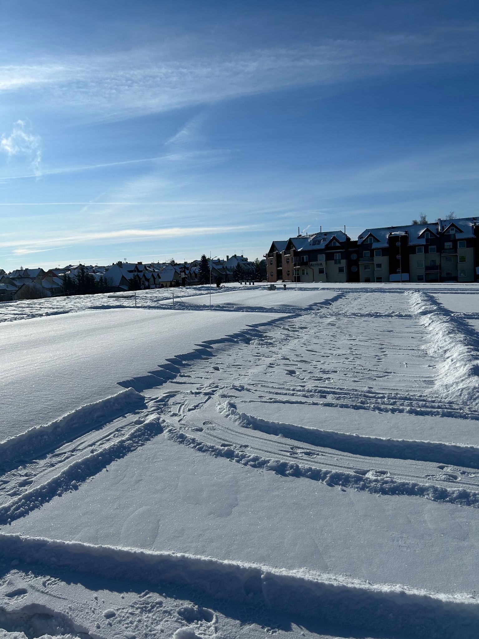 Śnieżny plac z odśnieżonymi pasami, widoczne budynki w tle pod błękitnym niebem. Ślady opon na śniegu sugerują niedawne prace.