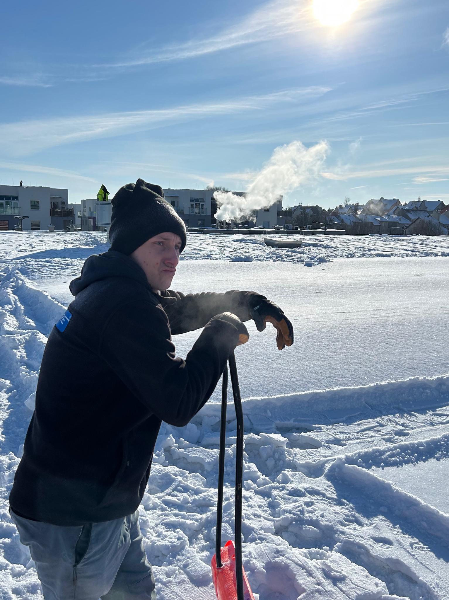 Mężczyzna w czapce i rękawicach opiera się na czerwonej łopacie do śniegu na zasypanym śniegiem placu, w tle budynki i para unosząca się z kominów.