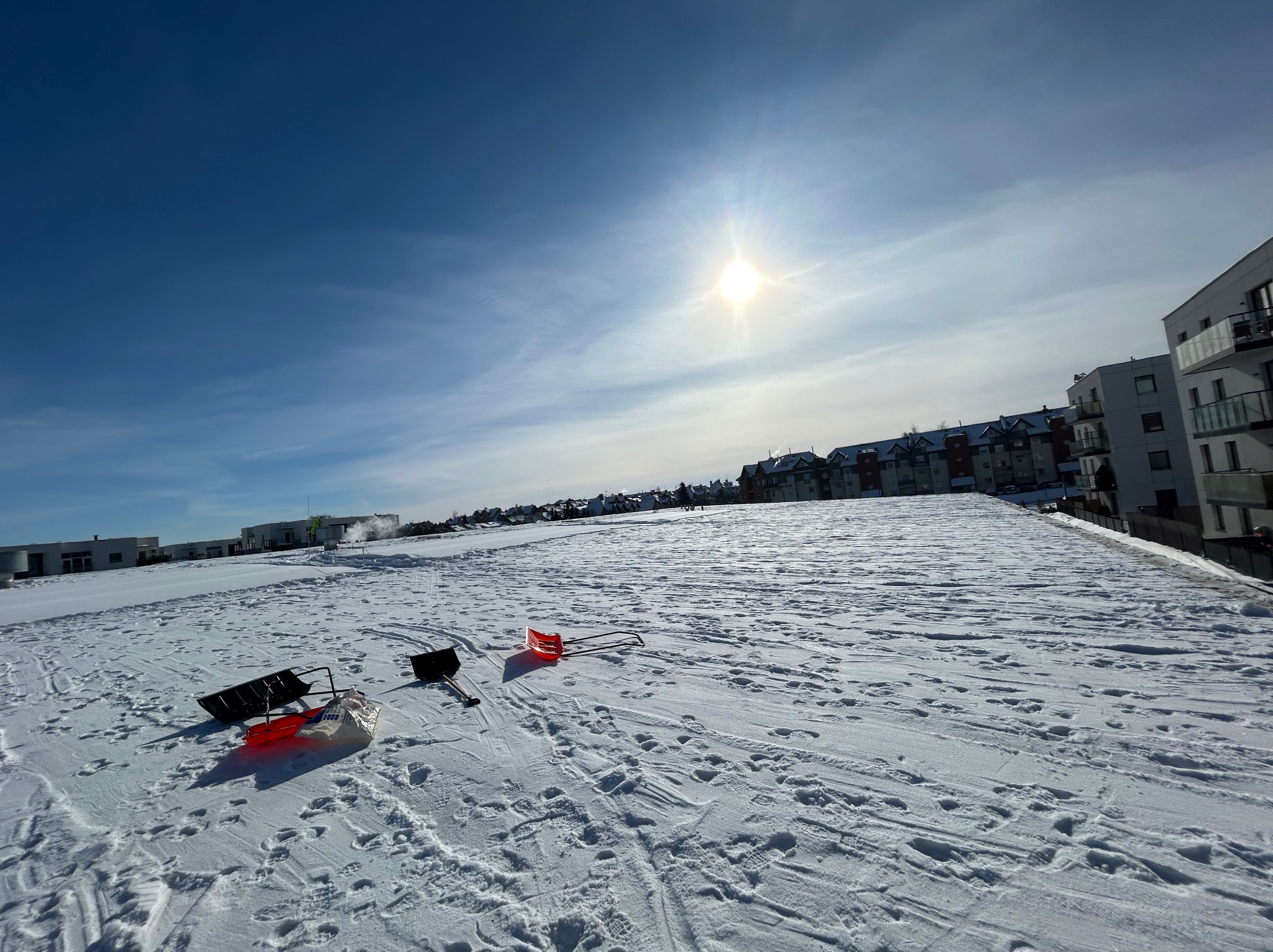 Śnieżny teren z narzędziami do odśnieżania: łopaty i sanki. W tle budynki mieszkalne pod błękitnym niebem z jasnym słońcem. Widoczne ślady na śniegu.