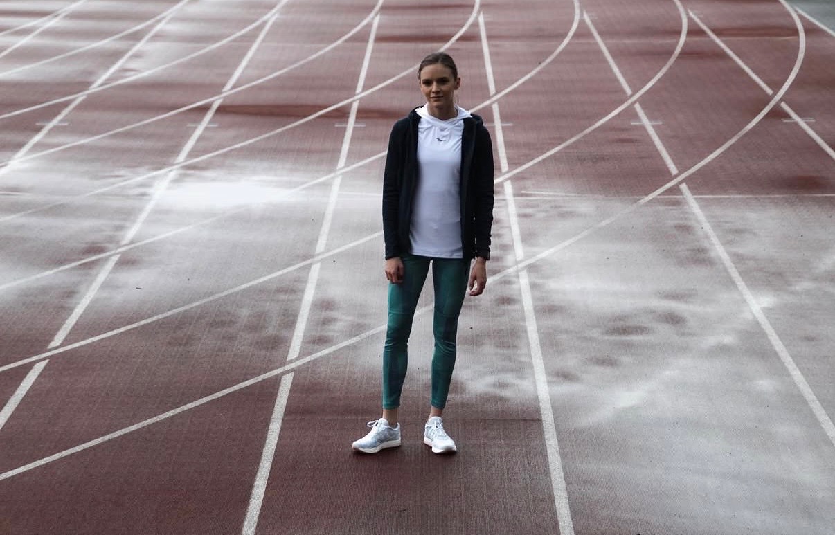 Kobieta w sportowym stroju stoi na bieżni lekkoatletycznej. Ubrana w bluzę, legginsy i buty do biegania. Widok z przodu, centralnie umieszczona postać.