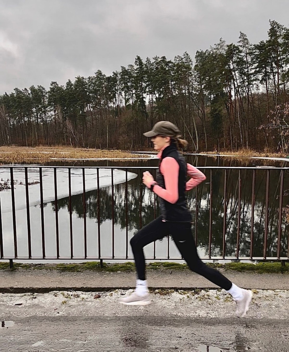 Kobieta w czarnym stroju biegowym i czapce, w dynamicznym biegu na tle jeziora i lasu w pochmurny dzień. Widok z boku, uchwycony w ruchu.