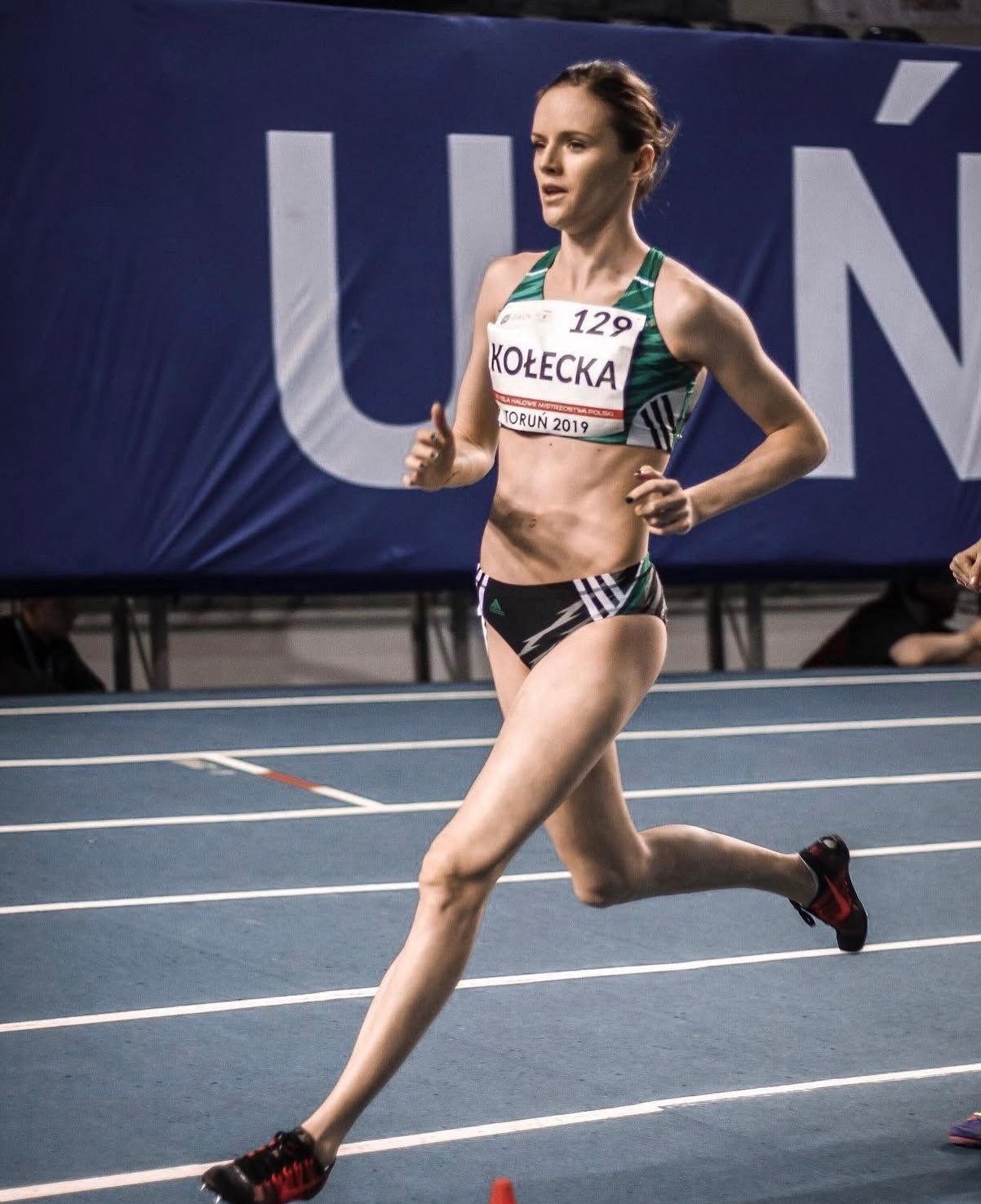 Kobieta-lekkoatletka w trakcie biegu na bieżni, w stroju sportowym z numerem 129, widoczny napis KOŁECKA i TORUŃ 2019. Dynamiczne ujęcie.