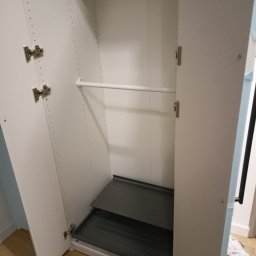 Montaż mebli paczkowanych Usługi transportowe - Wnętrze szafy z otwartymi drzwiami, widoczny drążek na ubrania i wysuwane półki. Białe płyty meblowe z licznymi otworami montażowymi.