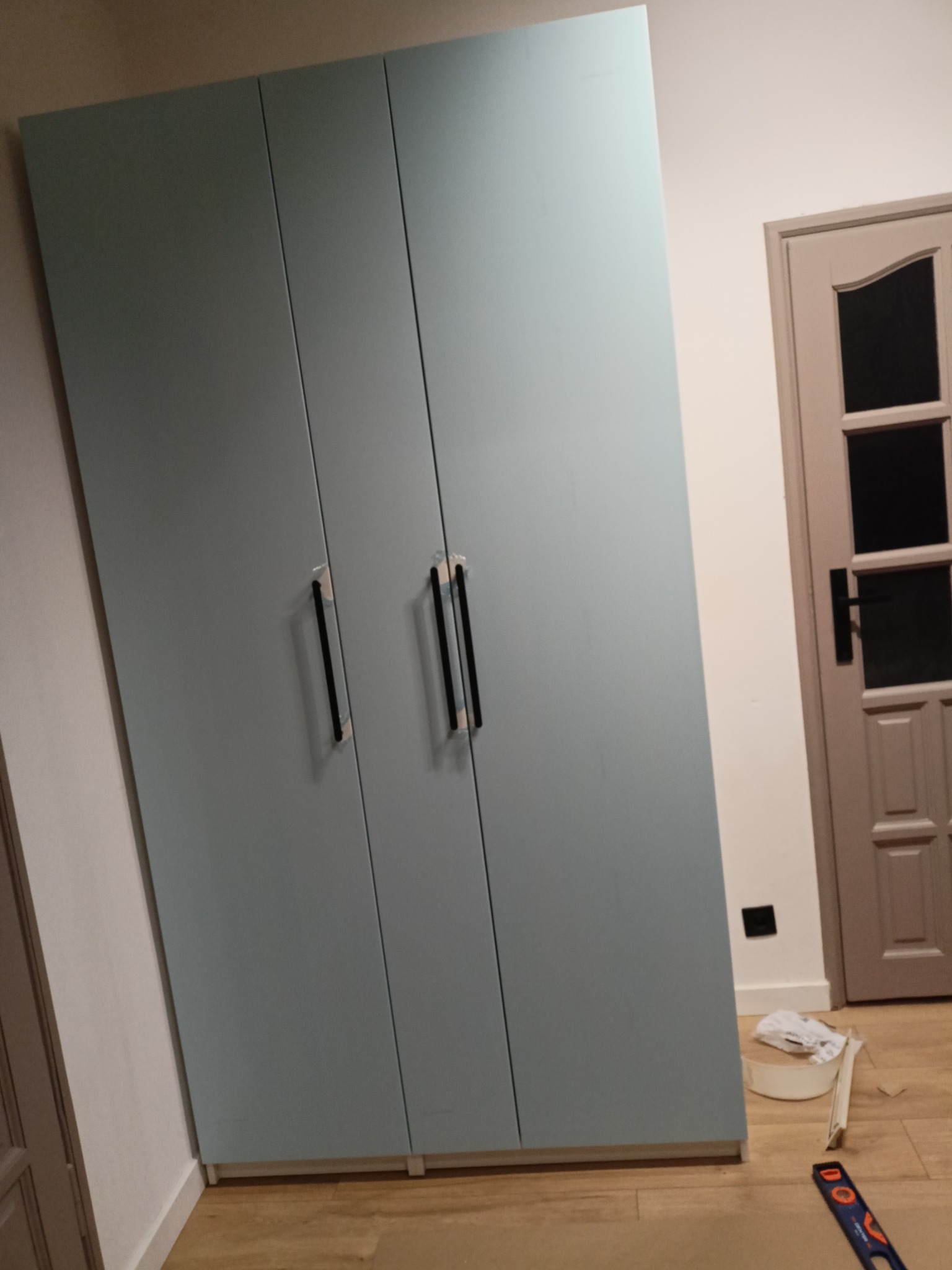 Jasnoniebieska szafa trzydrzwiowa z czarnymi uchwytami, ustawiona na drewnianej podłodze. Widoczne narzędzia i resztki materiałów po montażu. Drzwi w tle.