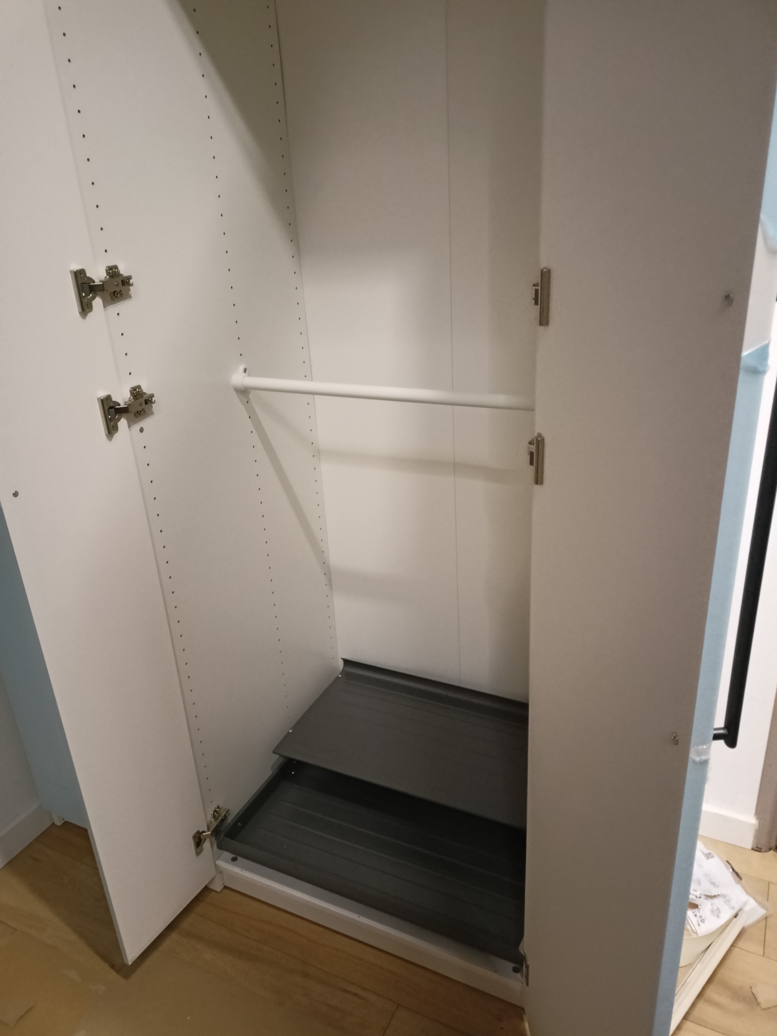 Wnętrze szafy z otwartymi drzwiami, widoczny drążek na ubrania i wysuwane półki. Białe płyty meblowe z licznymi otworami montażowymi.