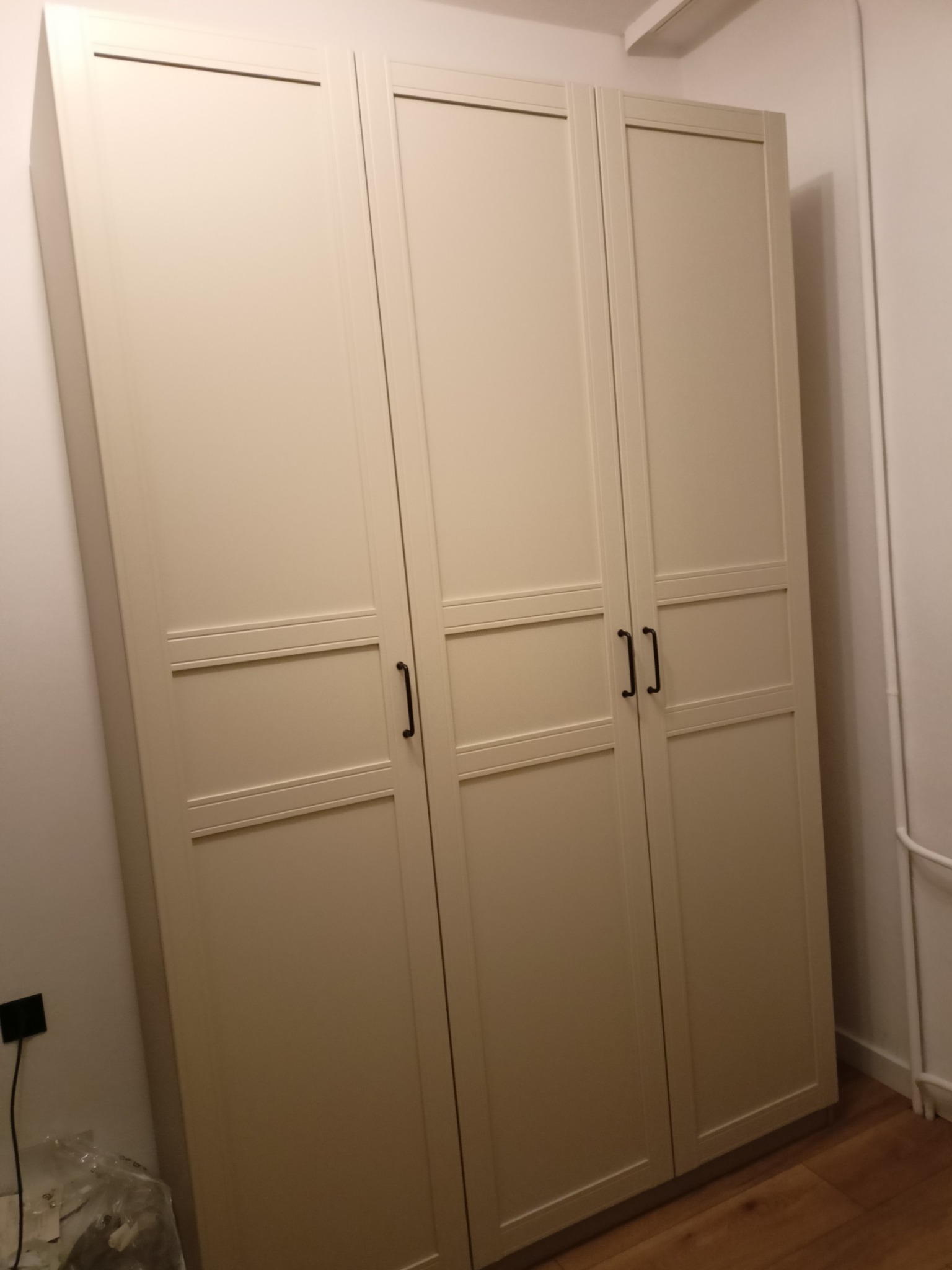 Zamknięta, beżowa szafa trzydrzwiowa z czarnymi uchwytami, ustawiona na drewnianej podłodze przy białej ścianie. Widoczny fragment listwy przypodłogowej i rury.