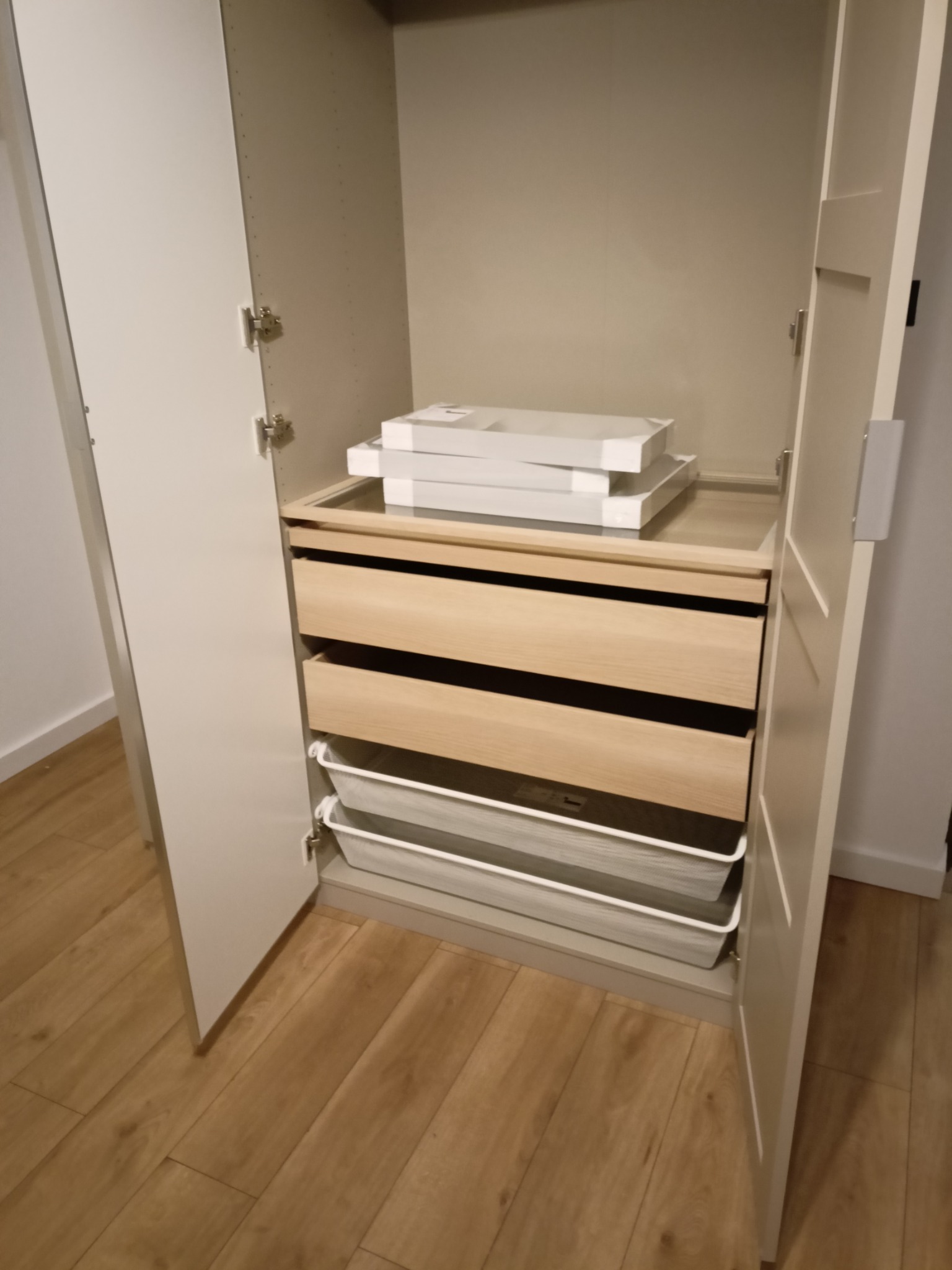 Wnętrze szafy z otwartymi drzwiami, widoczne drewniane szuflady, metalowe kosze i paczki na półce. Jasne kolory, minimalistyczny design.