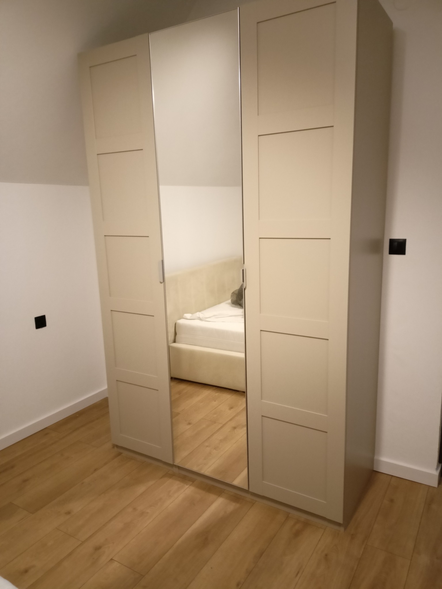 Beżowa szafa trzydrzwiowa z lustrem w sypialni na poddaszu. Widoczne drewniane panele podłogowe i fragment łóżka. Minimalistyczny design, neutralna kolorystyka.