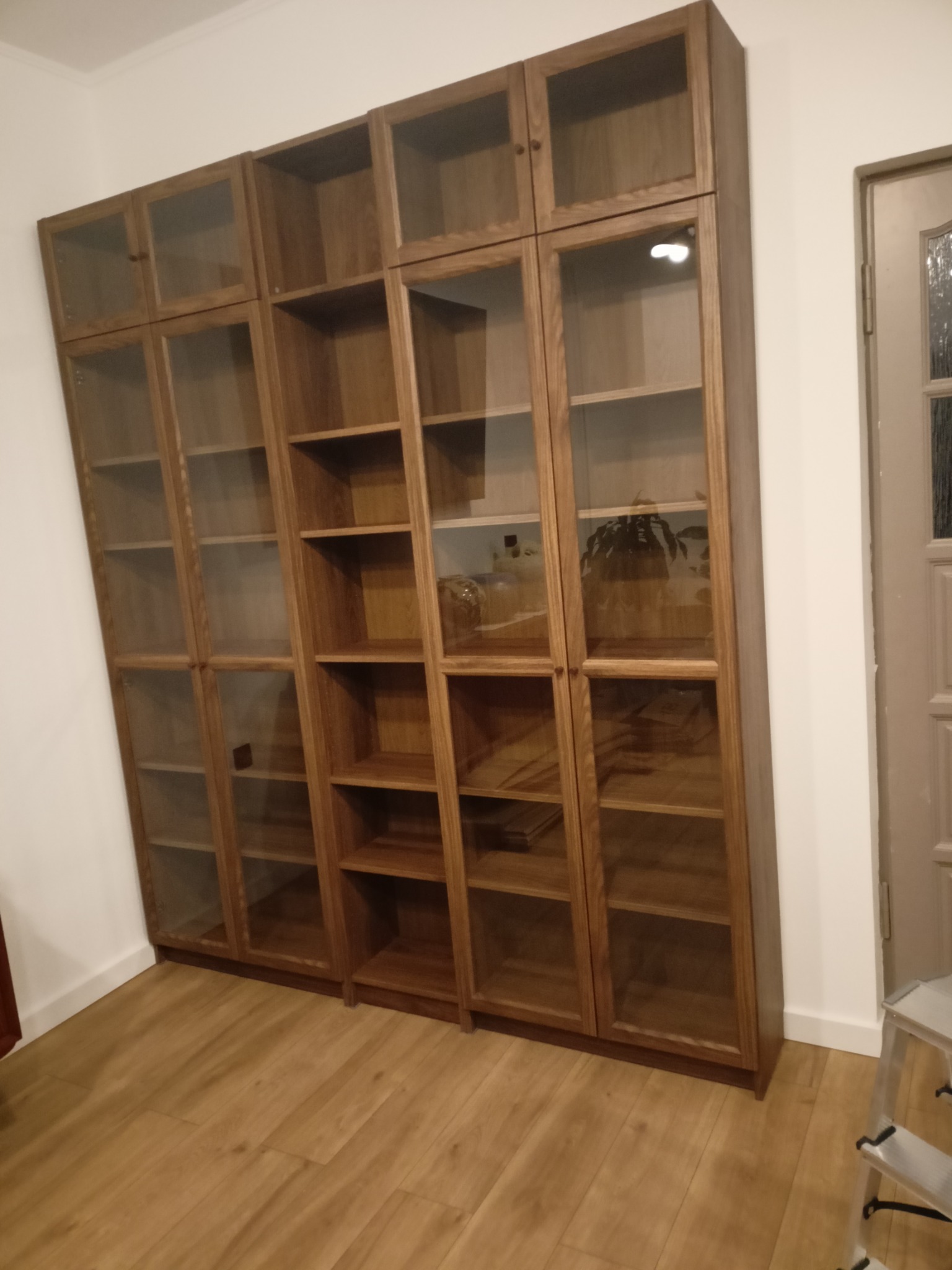 Drewniany regał z przeszklonymi drzwiczkami, podzielony na segmenty, ustawiony w rogu pokoju na drewnianej podłodze. Widoczna drabina malarska.