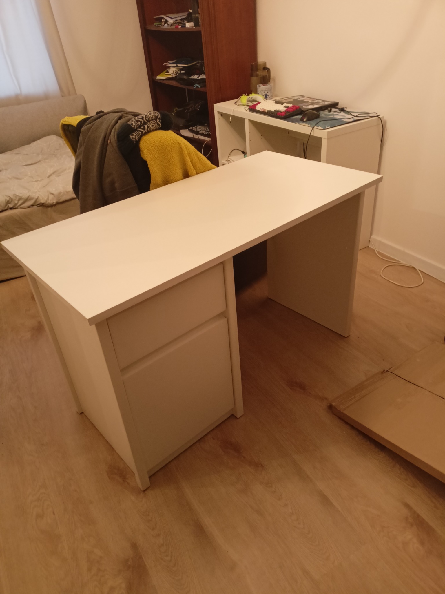 Białe biurko z szufladami, ustawione w pokoju na drewnianej podłodze. Na blacie brak przedmiotów, co podkreśla jego minimalistyczny design. Widoczny fragment łóżka i regału.