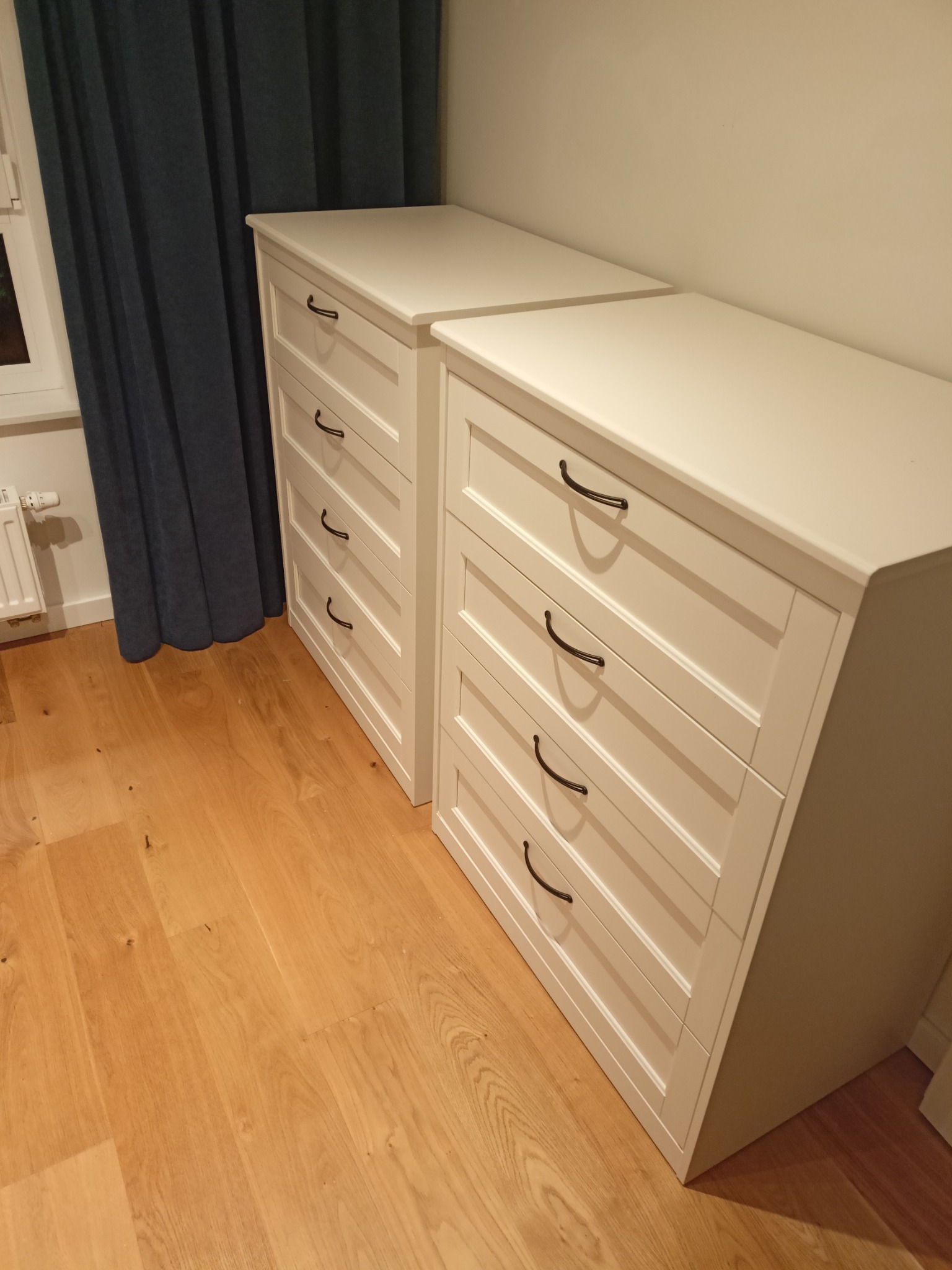 Dwie białe komody z czarnymi uchwytami na drewnianej podłodze, obok zasłony. Ustawione w rzędzie, tworzą spójną aranżację wnętrza.