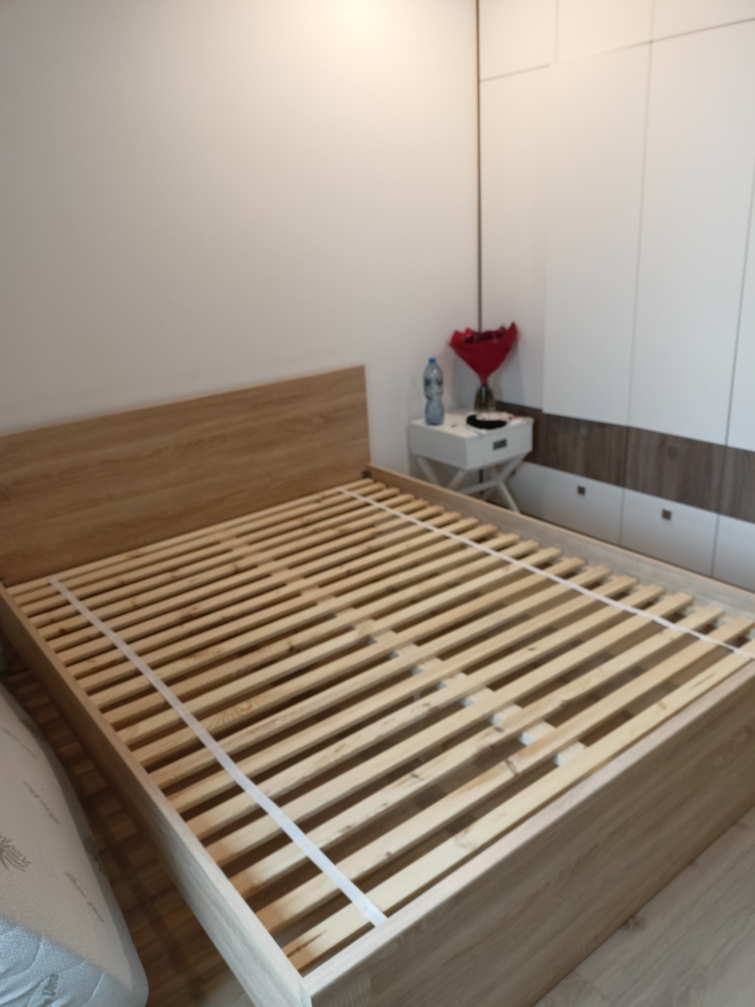 Drewniany stelaż łóżka z listewkami, częściowo zamontowany w ramie, widoczny w pokoju z szafą i stolikiem nocnym. Jasne wnętrze.