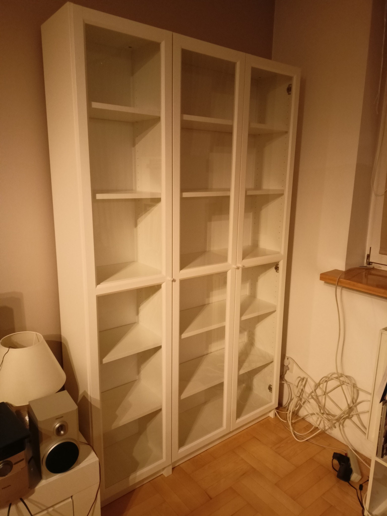 Biały regał z przeszklonymi drzwiami, trzy segmenty, widoczne półki, ustawiony na parkiecie w pokoju. U dołu widoczne kable i głośnik.