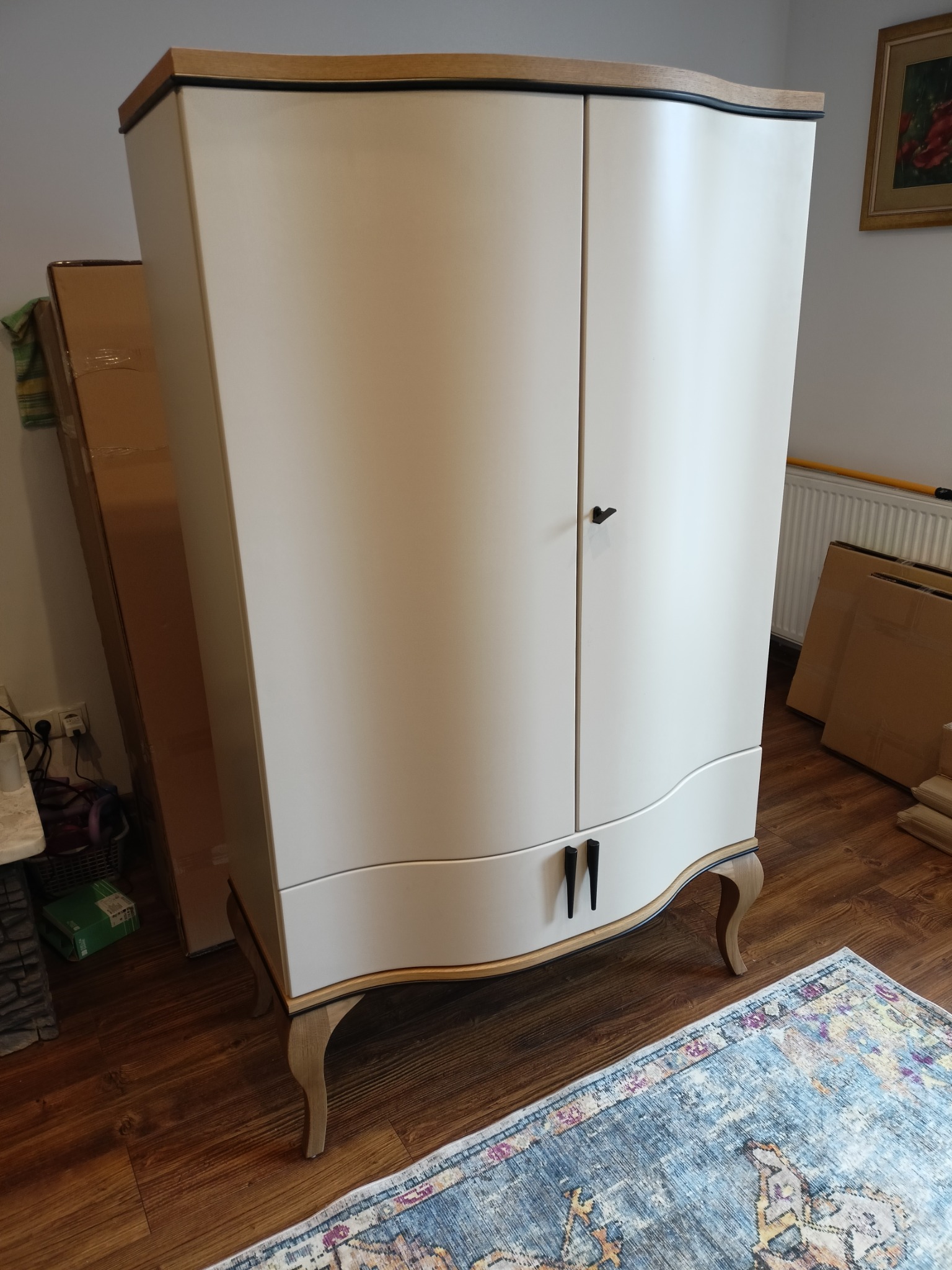 Dwudrzwiowa, kremowa szafa w stylu retro z drewnianymi akcentami i nóżkami, ustawiona na wzorzystym dywanie. W tle widoczne kartony i fragment ściany.