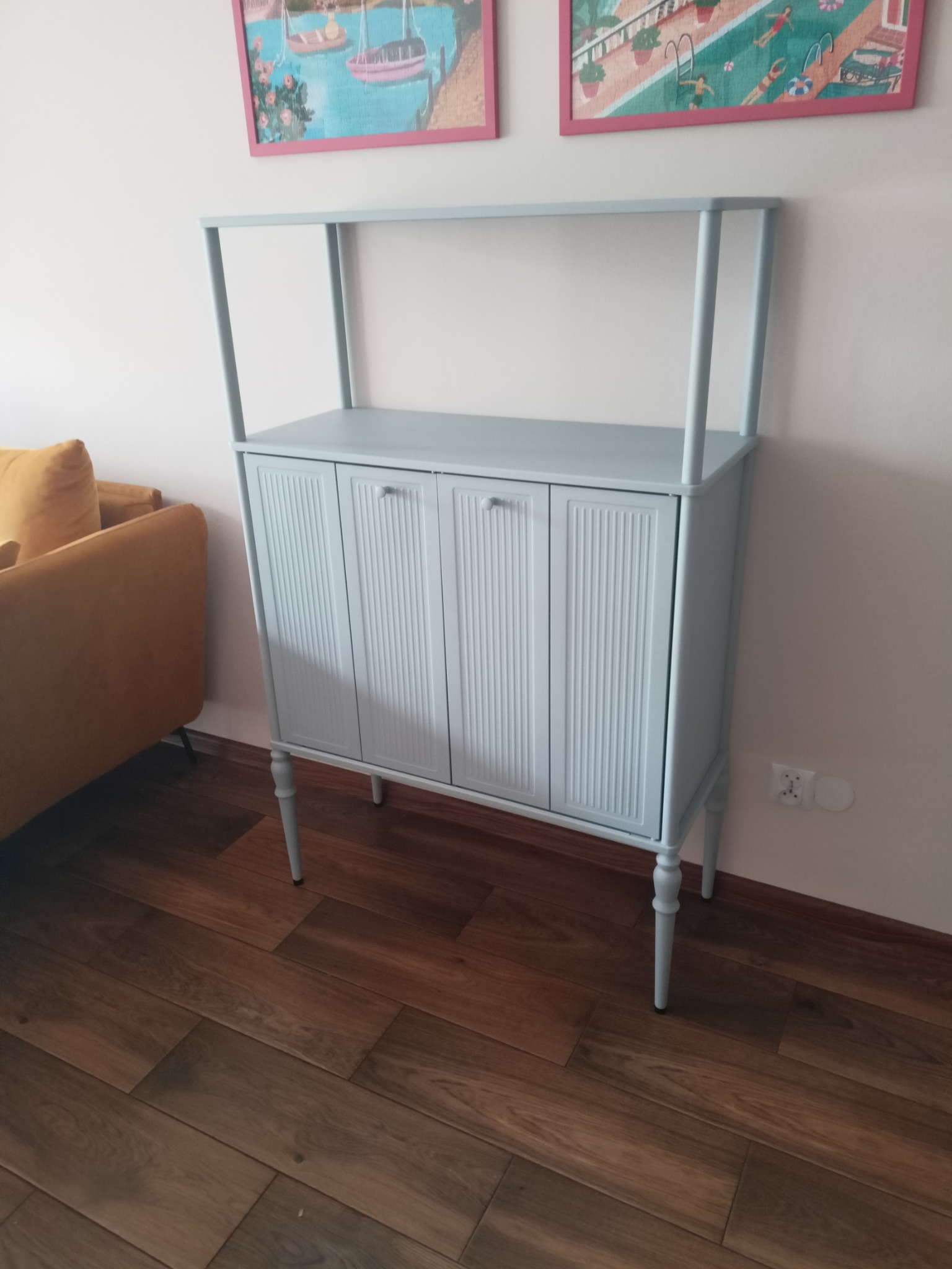 Jasnoniebieska komoda z trzema drzwiczkami i nóżkami w stylu retro, ustawiona na drewnianej podłodze, z fragmentem żółtej sofy w tle i obrazkami na ścianie.