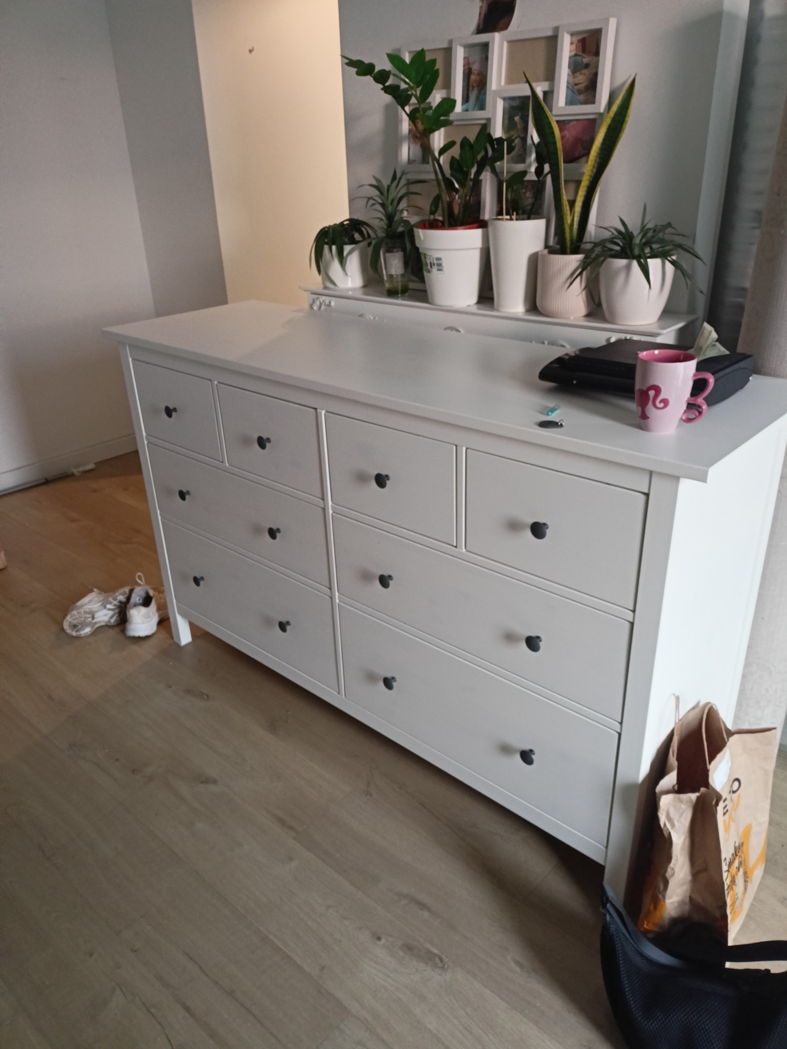 Biała komoda z czarnymi gałkami, na niej rośliny w doniczkach, obok torba i buty. Ustawiona na drewnianej podłodze w jasnym pokoju.