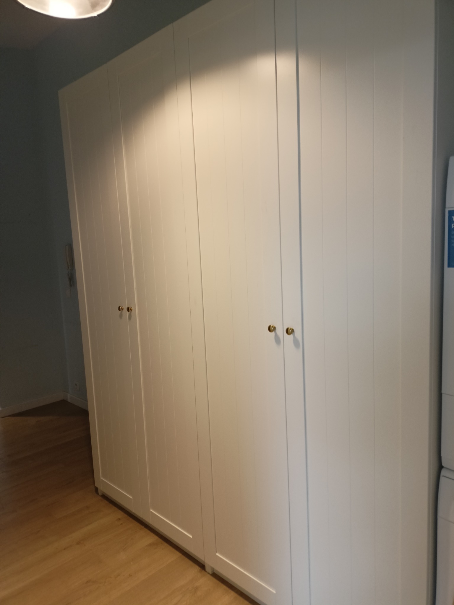 Biała, czterodrzwiowa szafa z pionowymi frezami i złotymi gałkami, ustawiona na drewnianej podłodze w jasnym wnętrzu. Widoczny fragment ściany i sufit z lampą.