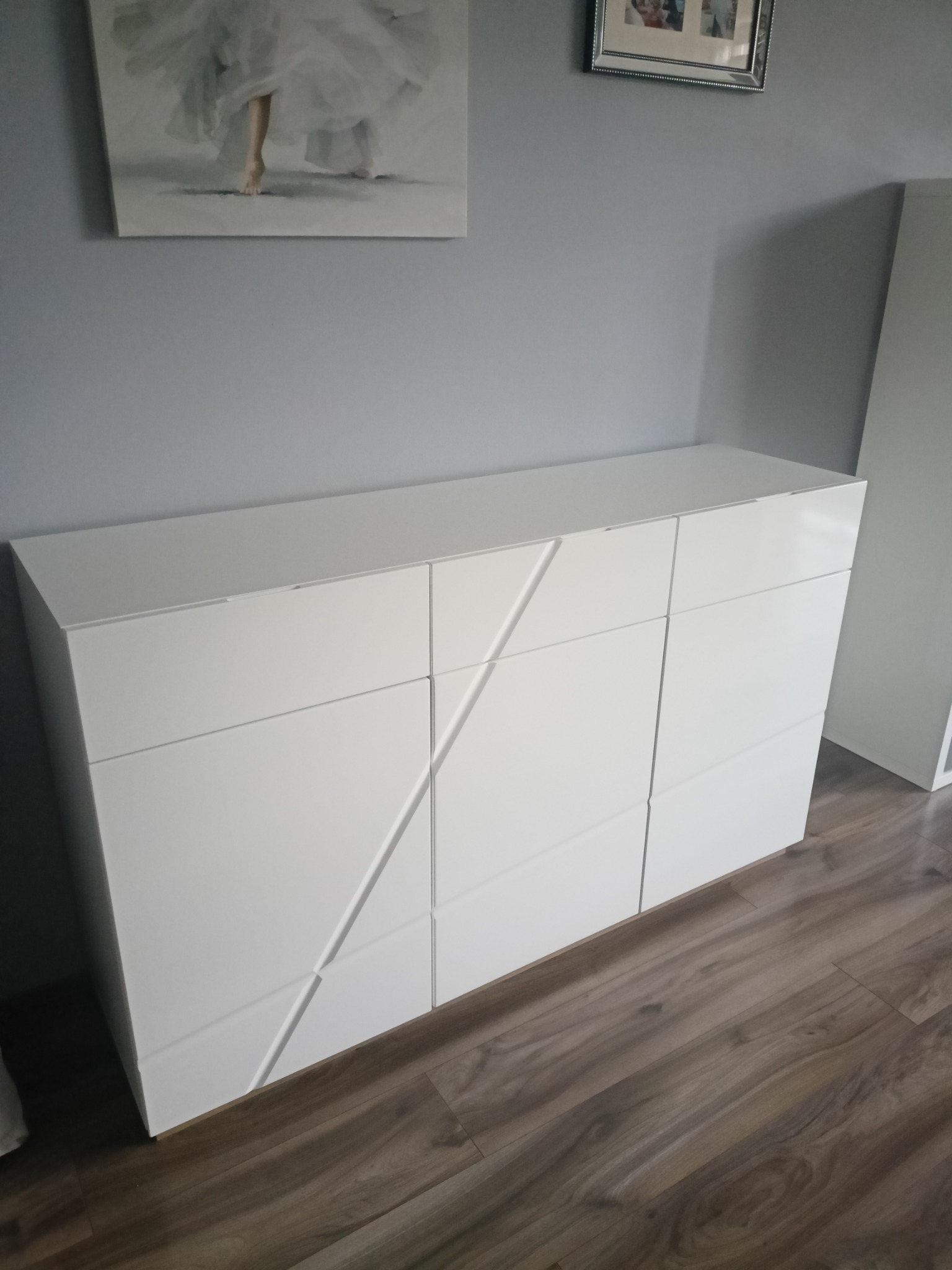 Biała komoda z sześcioma szufladami o geometrycznym wzorze na tle szarej ściany z obrazem i drewnianej podłogi. Nowoczesny design.