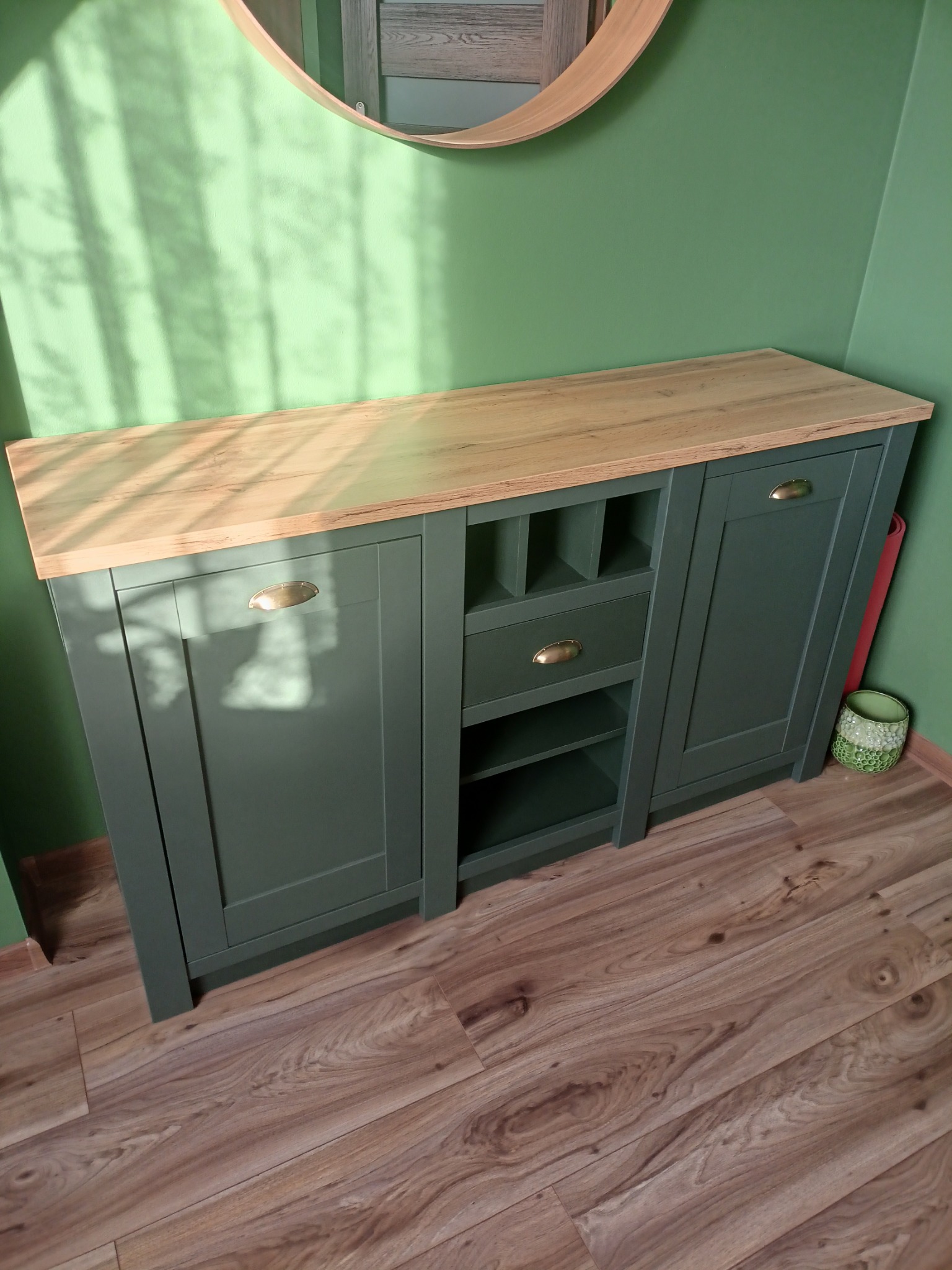 Zielony kredens z drewnianym blatem, złote uchwyty, okrągłe lustro w tle. Ustawiony na drewnianej podłodze, oświetlony promieniami słońca.