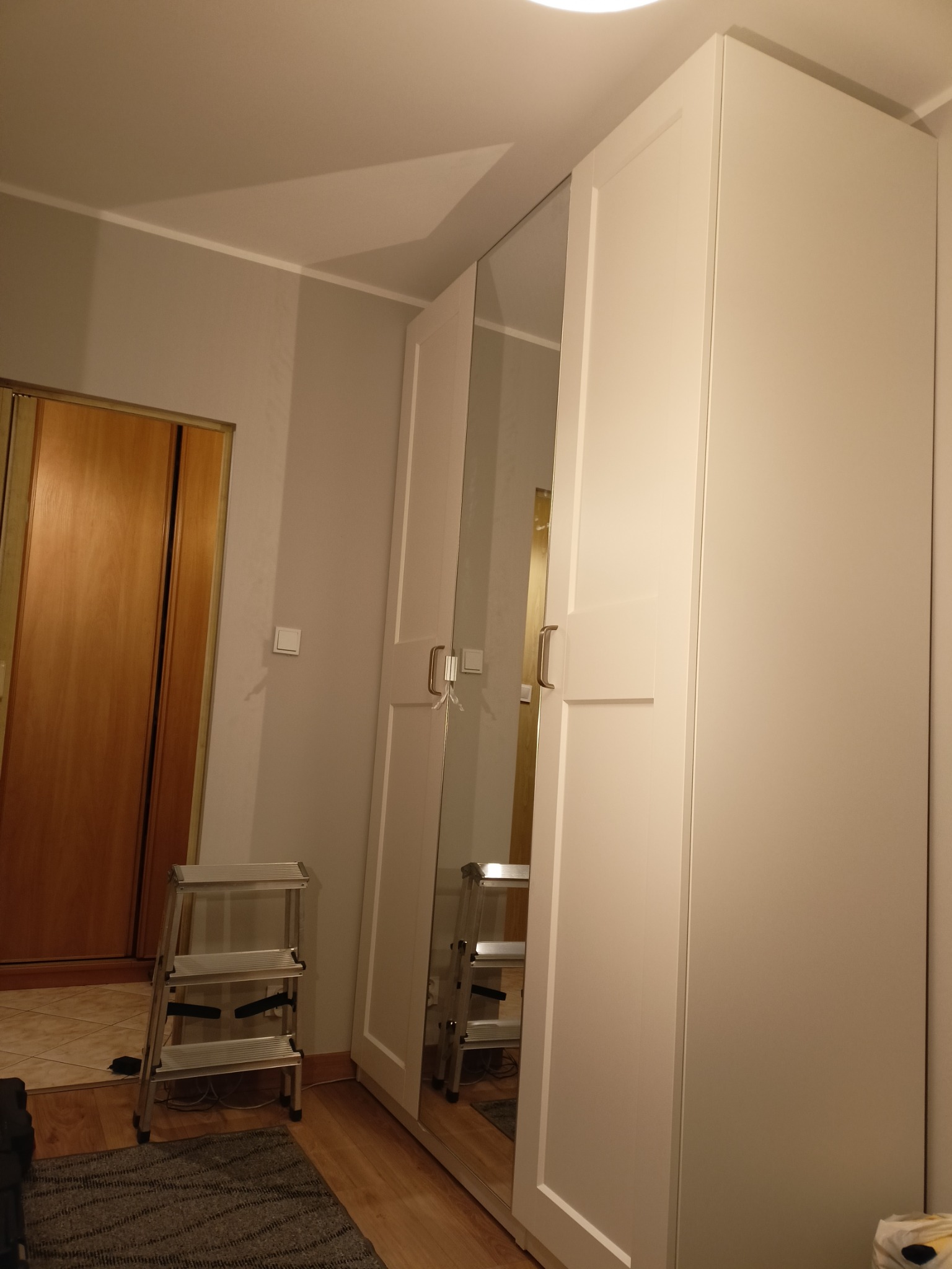 Biała, trzydrzwiowa szafa z lustrem, ustawiona w rogu pokoju. Obok drabina, widoczna część dywanu i drzwi do innego pomieszczenia. Ujęcie z dołu.