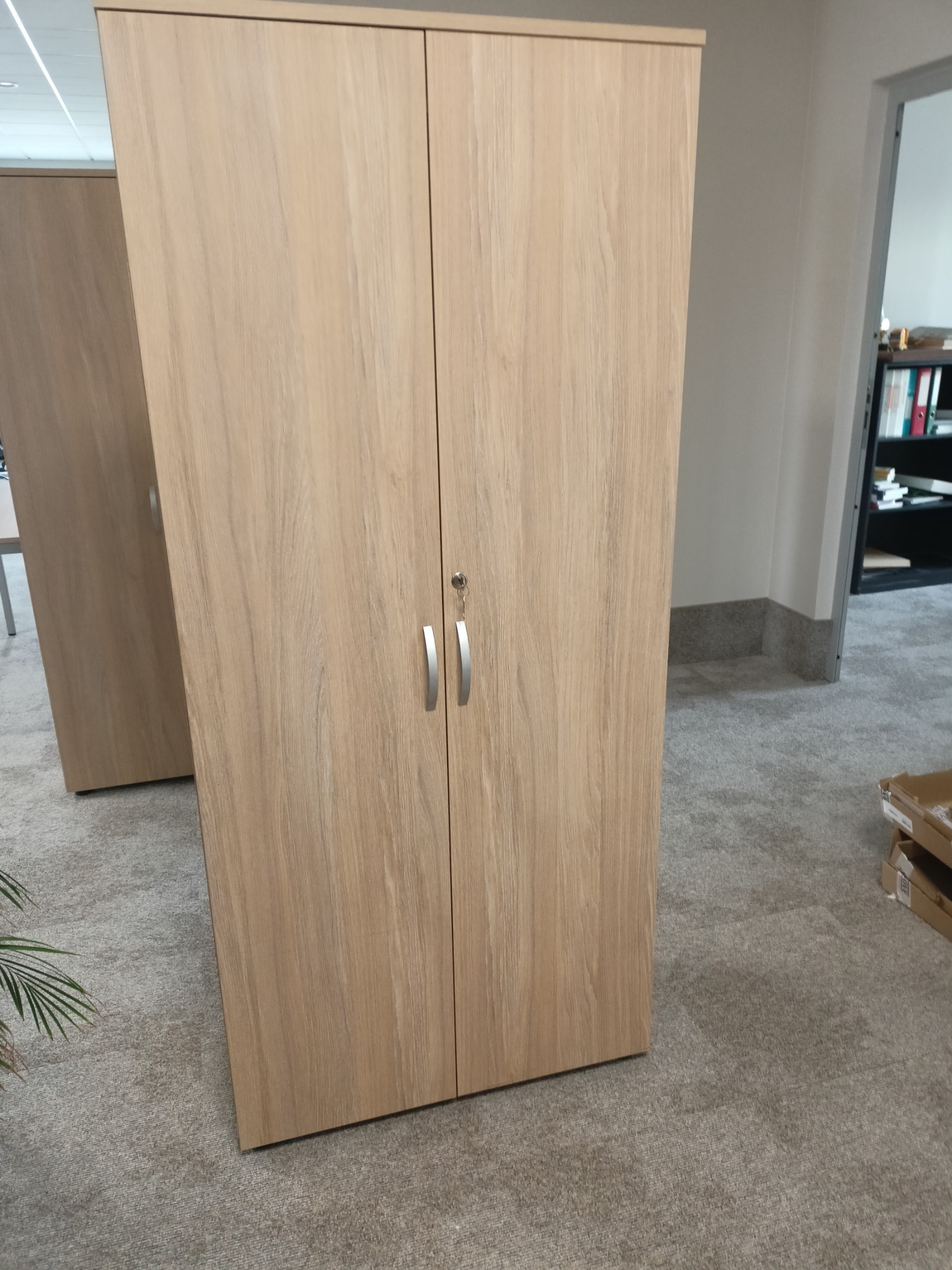Dwudrzwiowa szafa z jasnego drewna z metalowymi uchwytami, widoczna w biurze na tle szarej wykładziny i regału z segregatorami. Klucz w zamku.