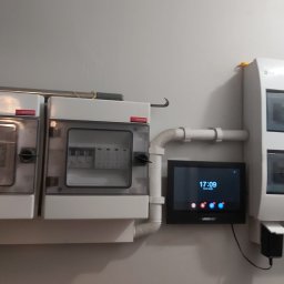 Pawel Jarocha - Dwa białe rozdzielnice elektryczne z przezroczystymi drzwiczkami, połączone rurką, obok czarny panel sterowania na szarej ścianie. Widoczne bezpieczniki i przewody.