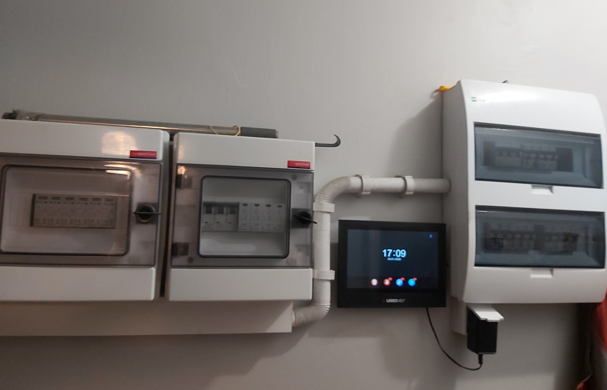 Dwa białe rozdzielnice elektryczne z przezroczystymi drzwiczkami, połączone rurką, obok czarny panel sterowania na szarej ścianie. Widoczne bezpieczniki i przewody.