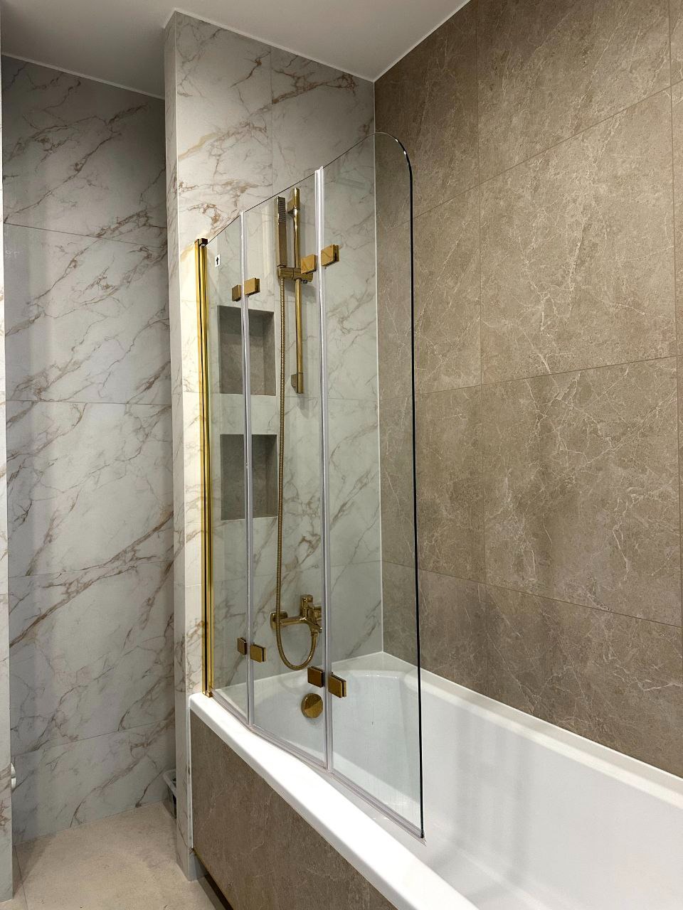 Wykończona łazienka z wanną i szklanym parawanem w złotych ramach. Marmurowe płytki na ścianie, nowoczesna armatura. Widok na detale wykończenia.