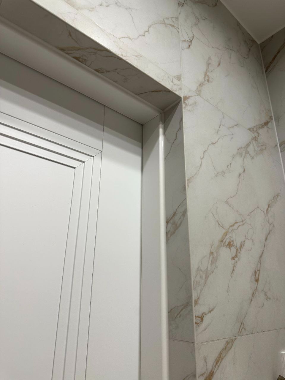 Białe drzwi z ościeżnicą i marmurowe płytki ścienne z brązowymi żyłami. Detal wykończenia wnętrza, kątowe ujęcie.
